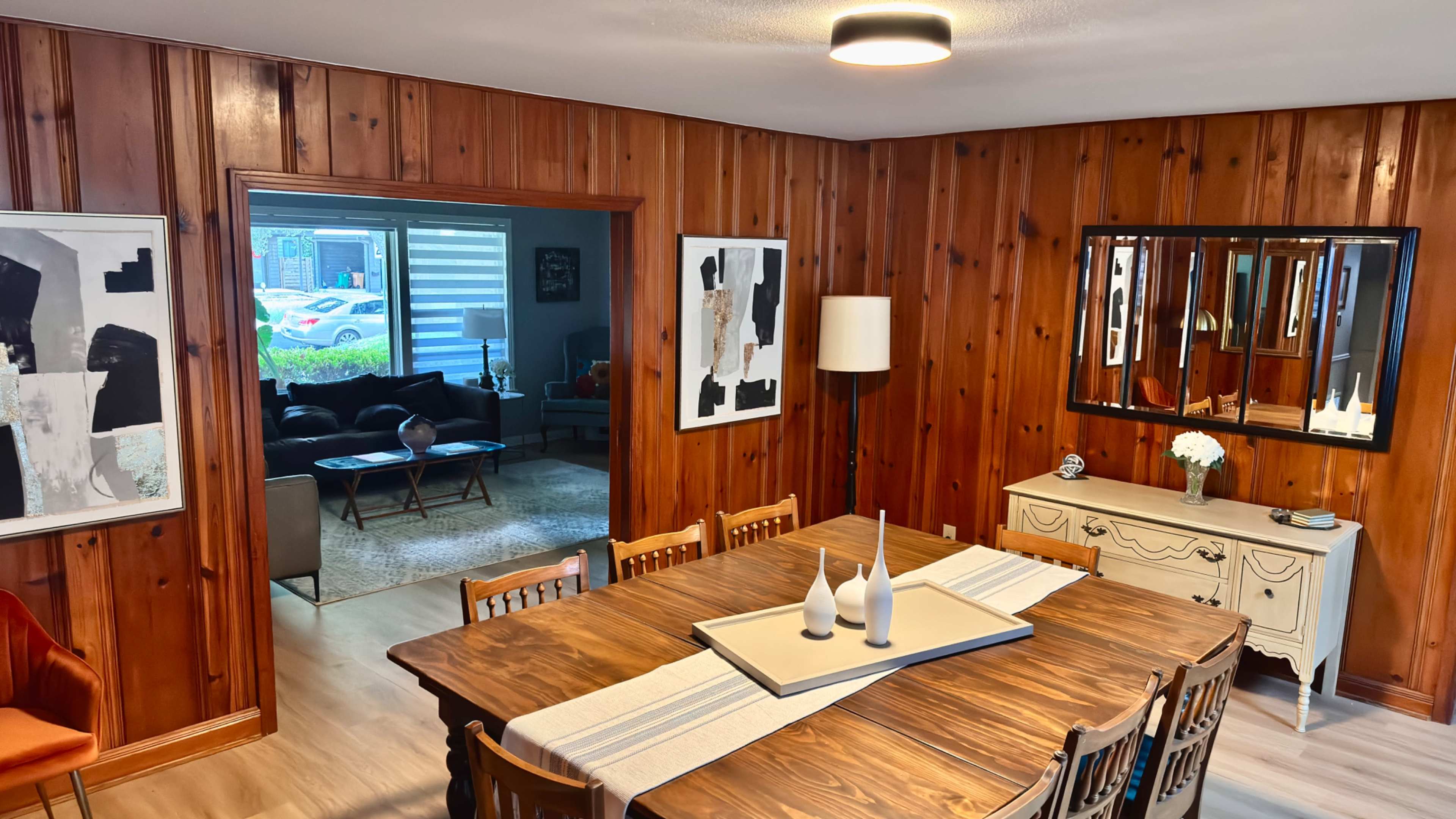 The image shows a wooden-paneled dining area with a large table and decorative vases, leading to an adjacent living space visible through an open doorway.