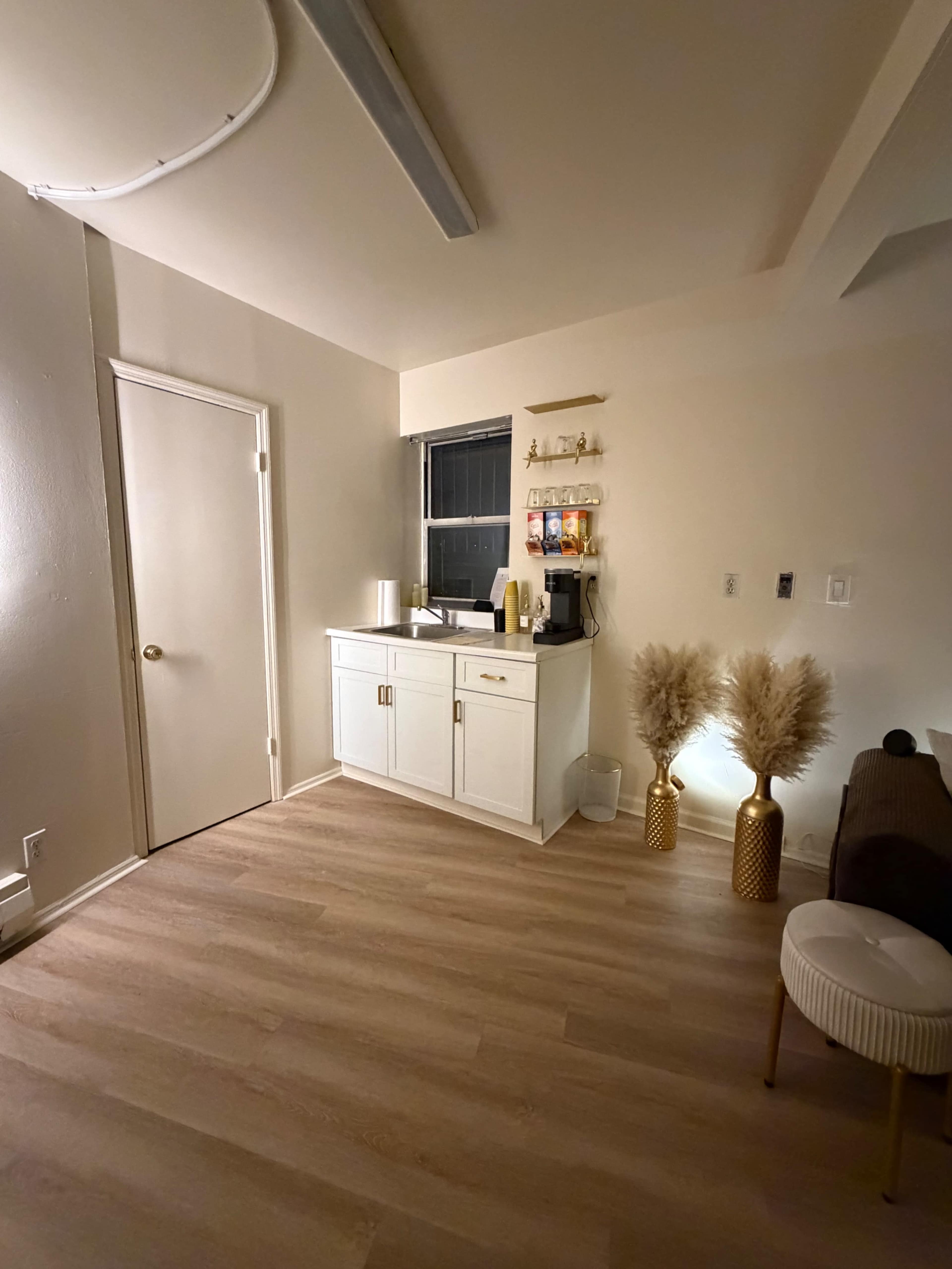 The image shows a cozy interior space featuring a white cabinet and a coffee station against a light-colored wall, with decorative plants in gold vases and a stylish couch.