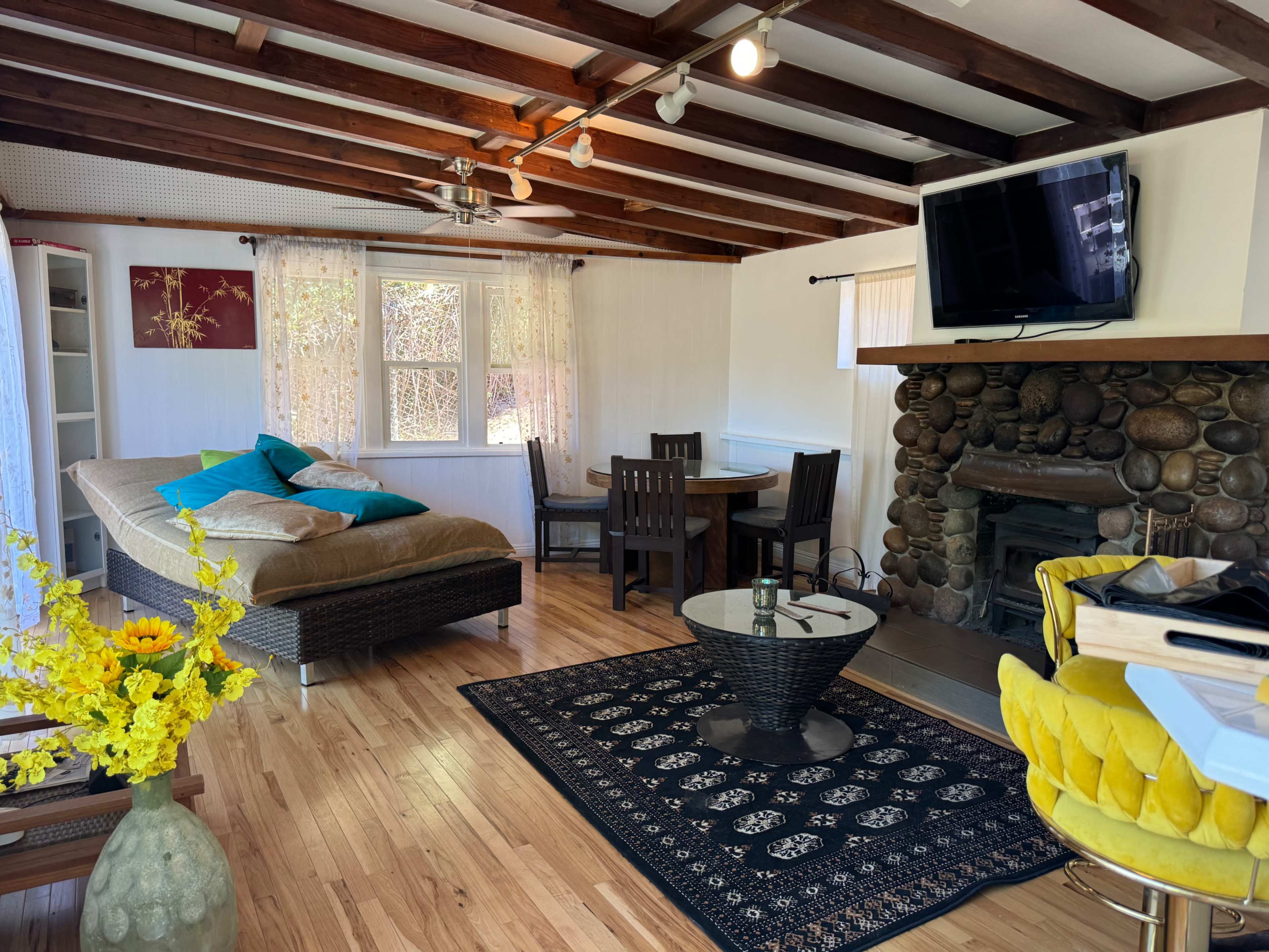 The image shows a cozy living space with wooden beams, a stone fireplace, a sofa, a dining table, and a yellow chair.