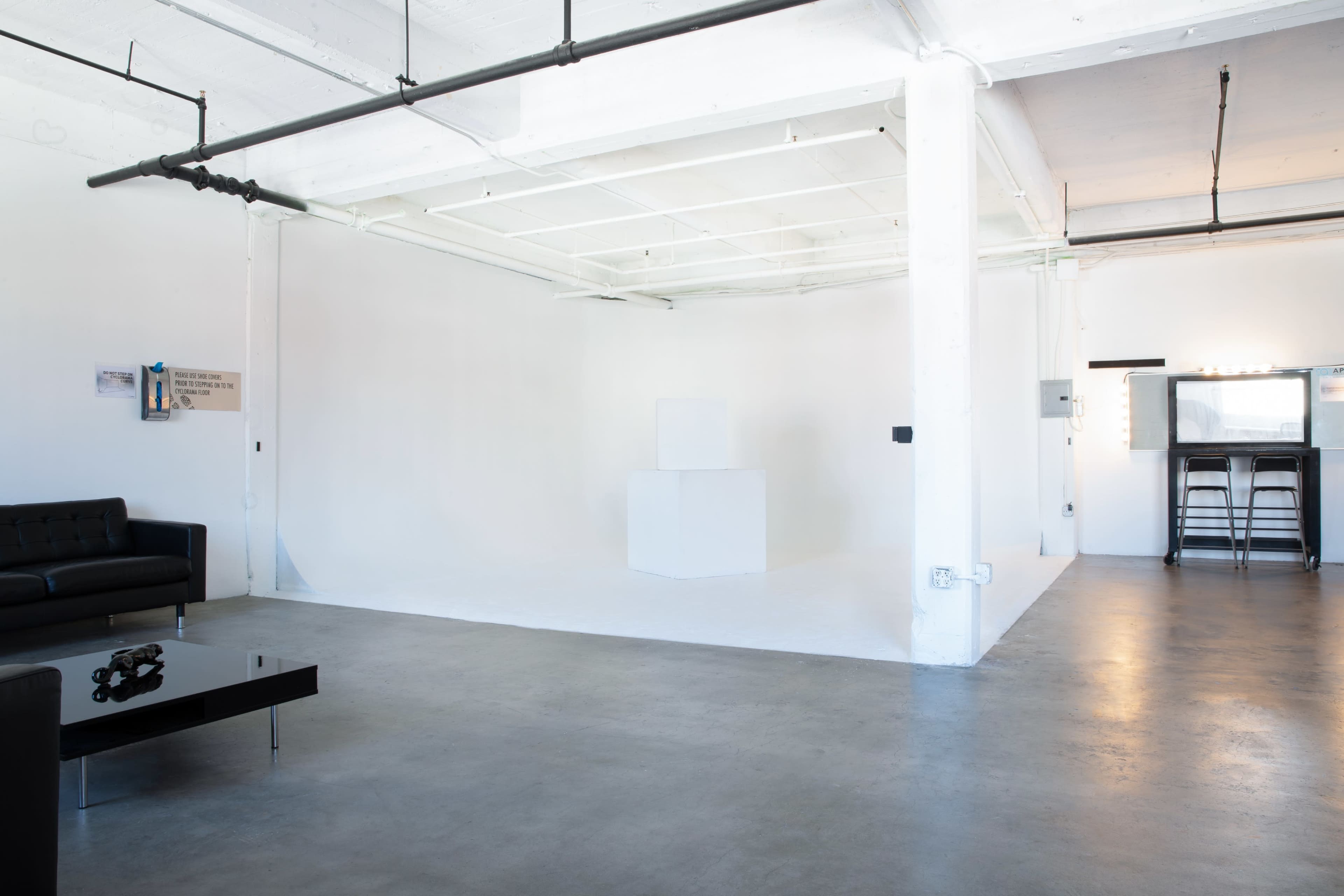 The image depicts a spacious, empty studio with white walls, a concrete floor, and minimal furnishings, including a black sofa and a coffee table.