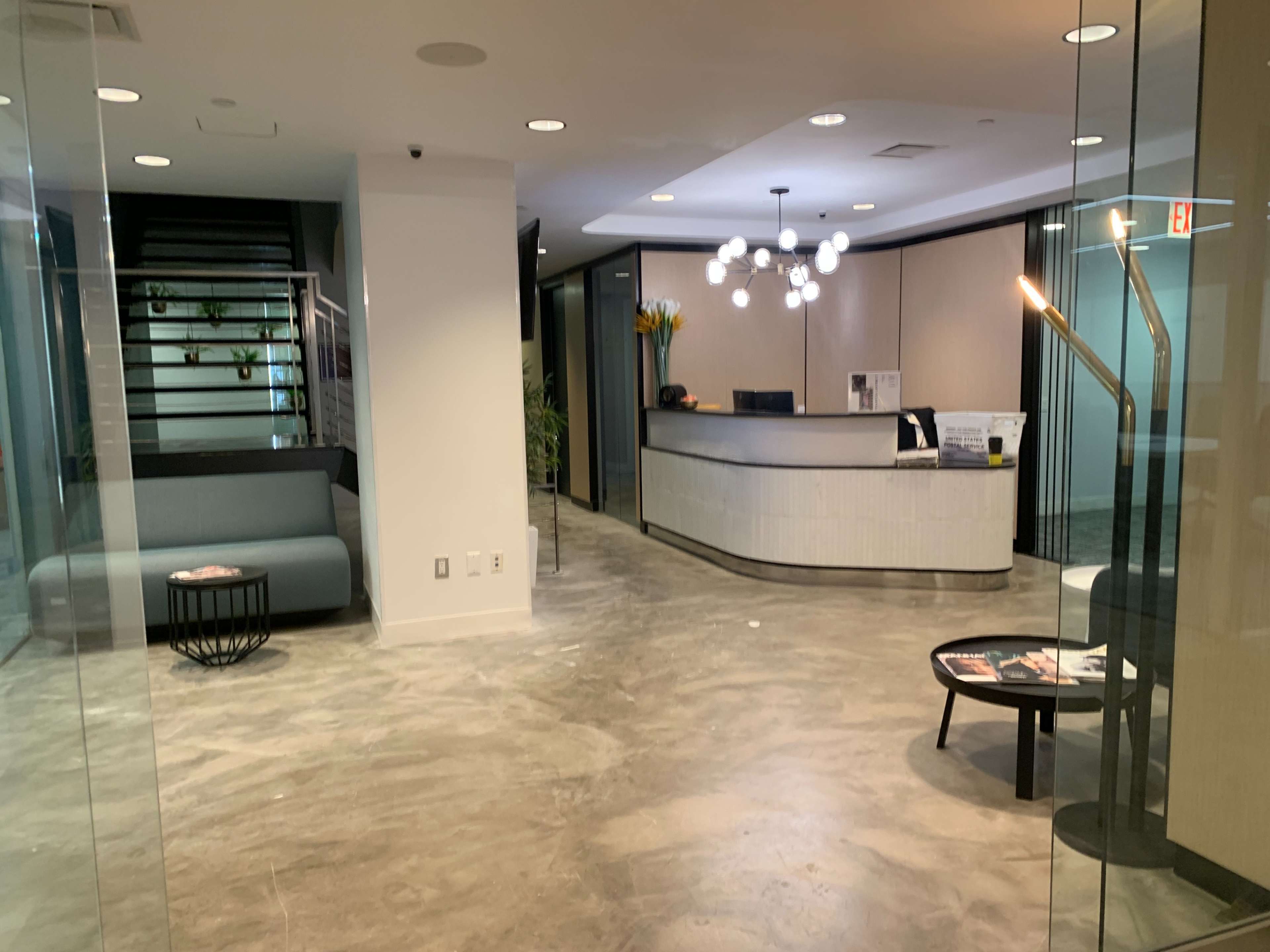 The image shows a modern reception area featuring a front desk, a seating area with a gray sofa, and contemporary lighting fixtures.