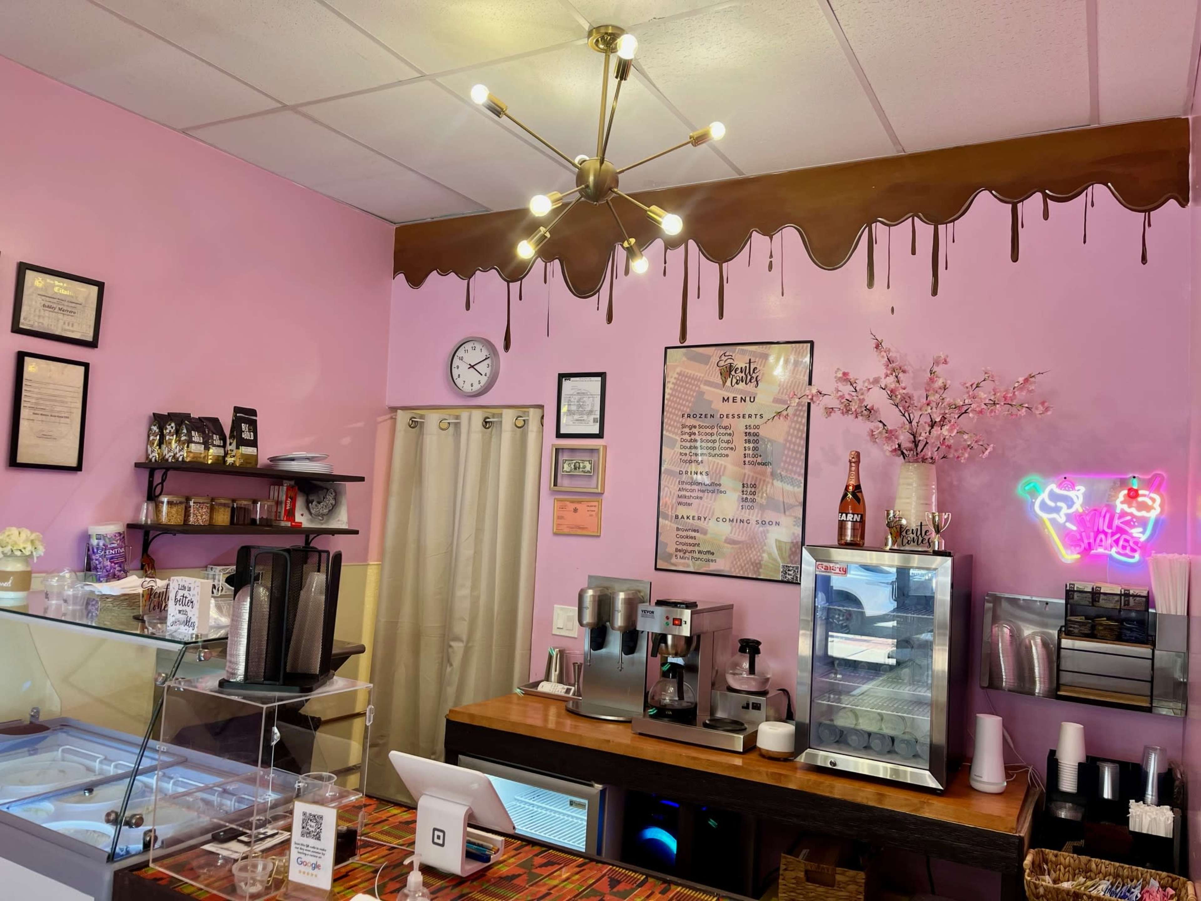 The image shows the interior of a cafe with pink walls, a display counter, and a menu board, featuring decorative elements like a chandelier and a neon sign.
