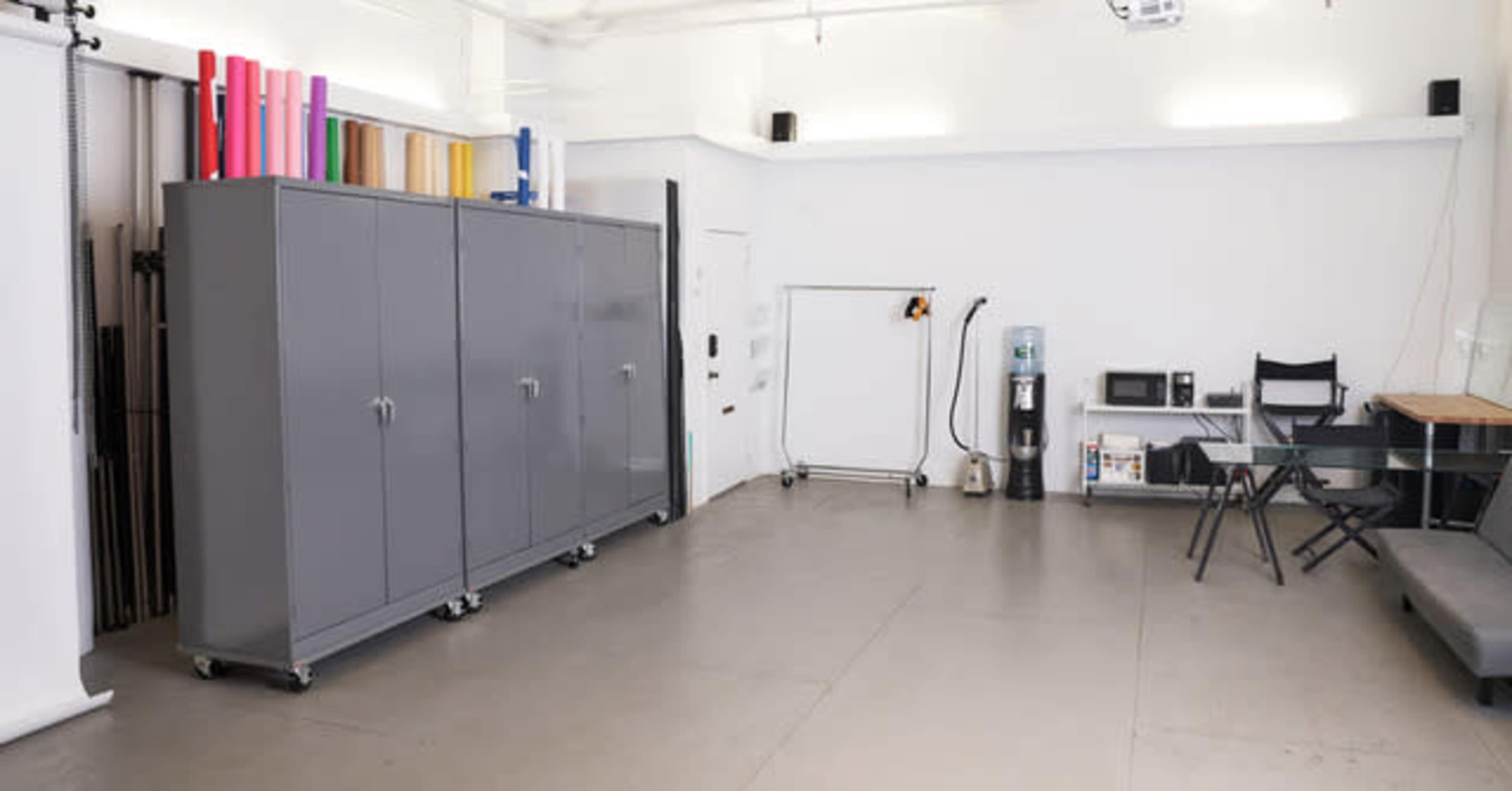 The image shows a spacious, well-organized studio with gray cabinets, a rolling garment rack, a microwave, and a table with chairs.