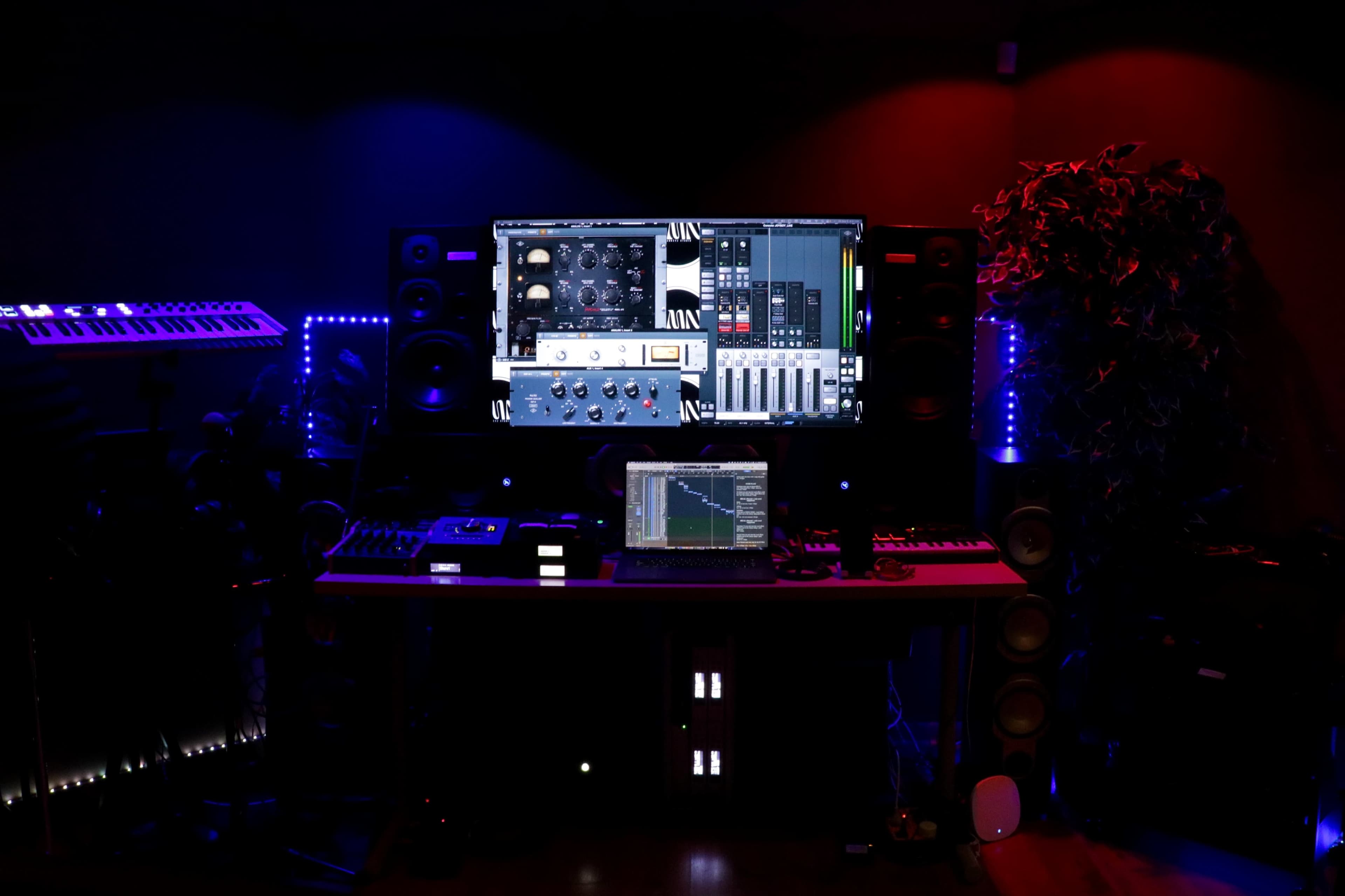 A music production studio is displayed with a large computer monitor, a laptop on a desk, various audio equipment, and ambient lighting in blue and red.