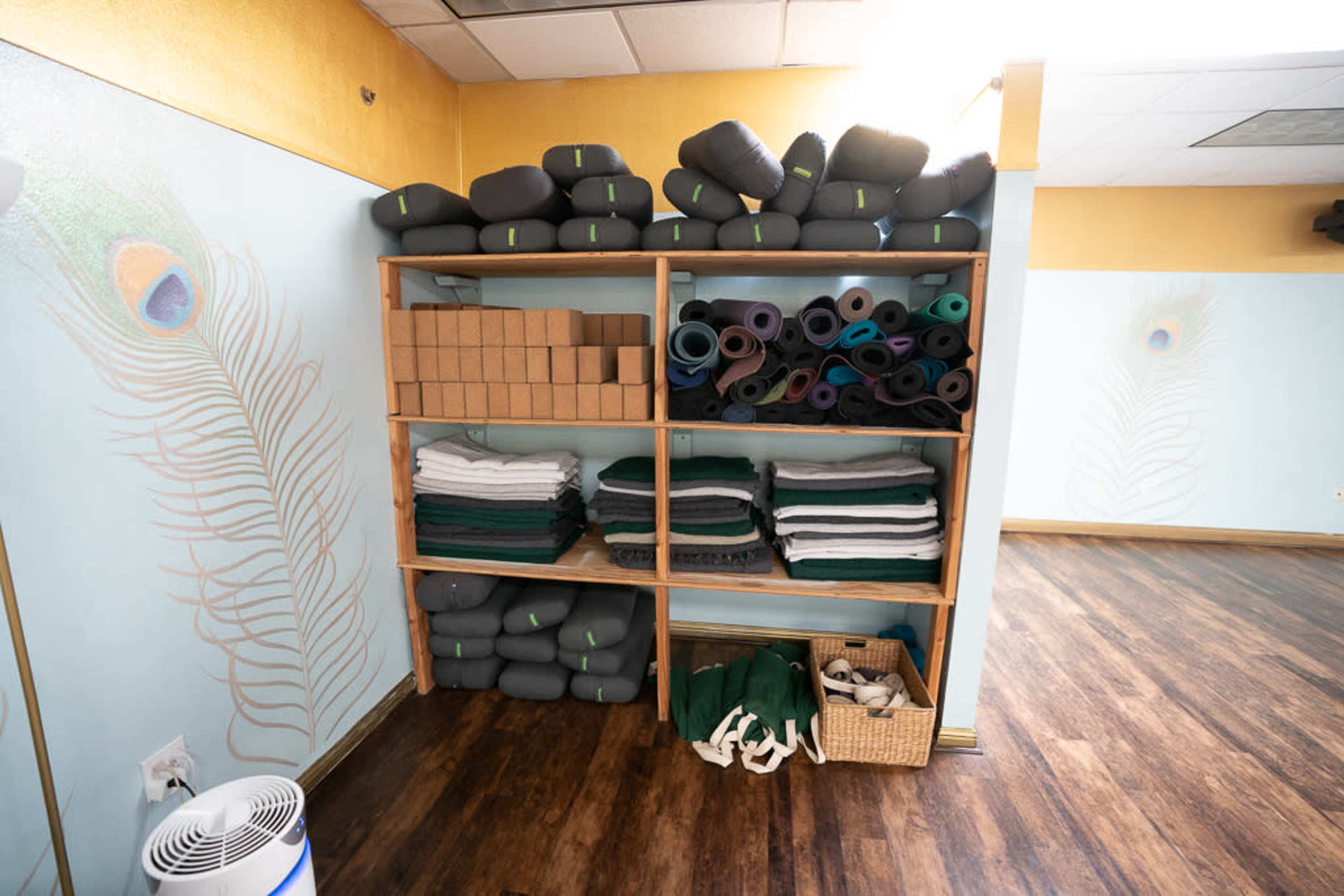 A wooden shelf holds neatly stacked yoga mats, blankets, and blocks in a light-filled studio with peacock feather decorations on the walls.