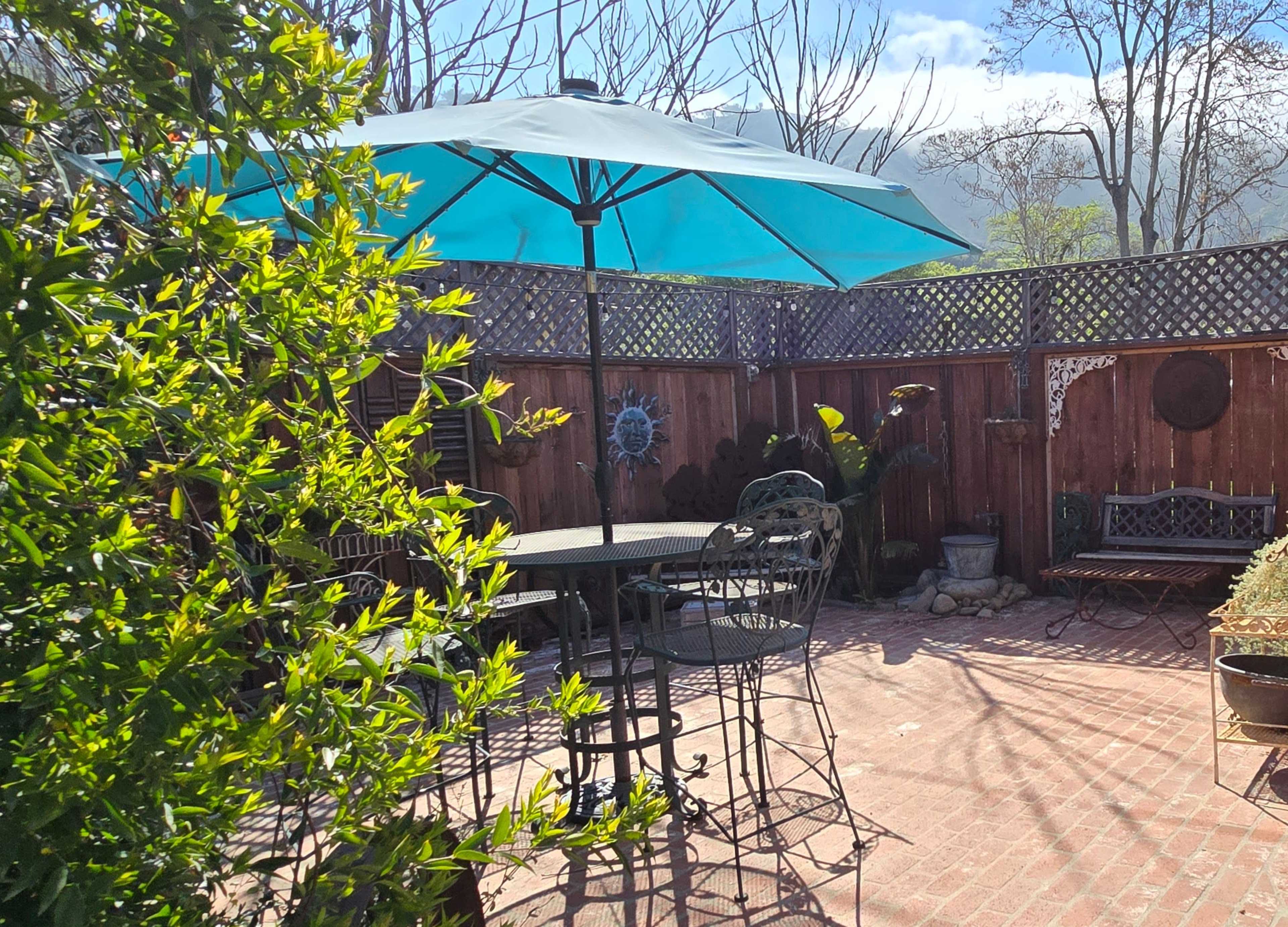 A patio area features a round table and chairs beneath a large turquoise umbrella, surrounded by greenery and a wooden fence.
