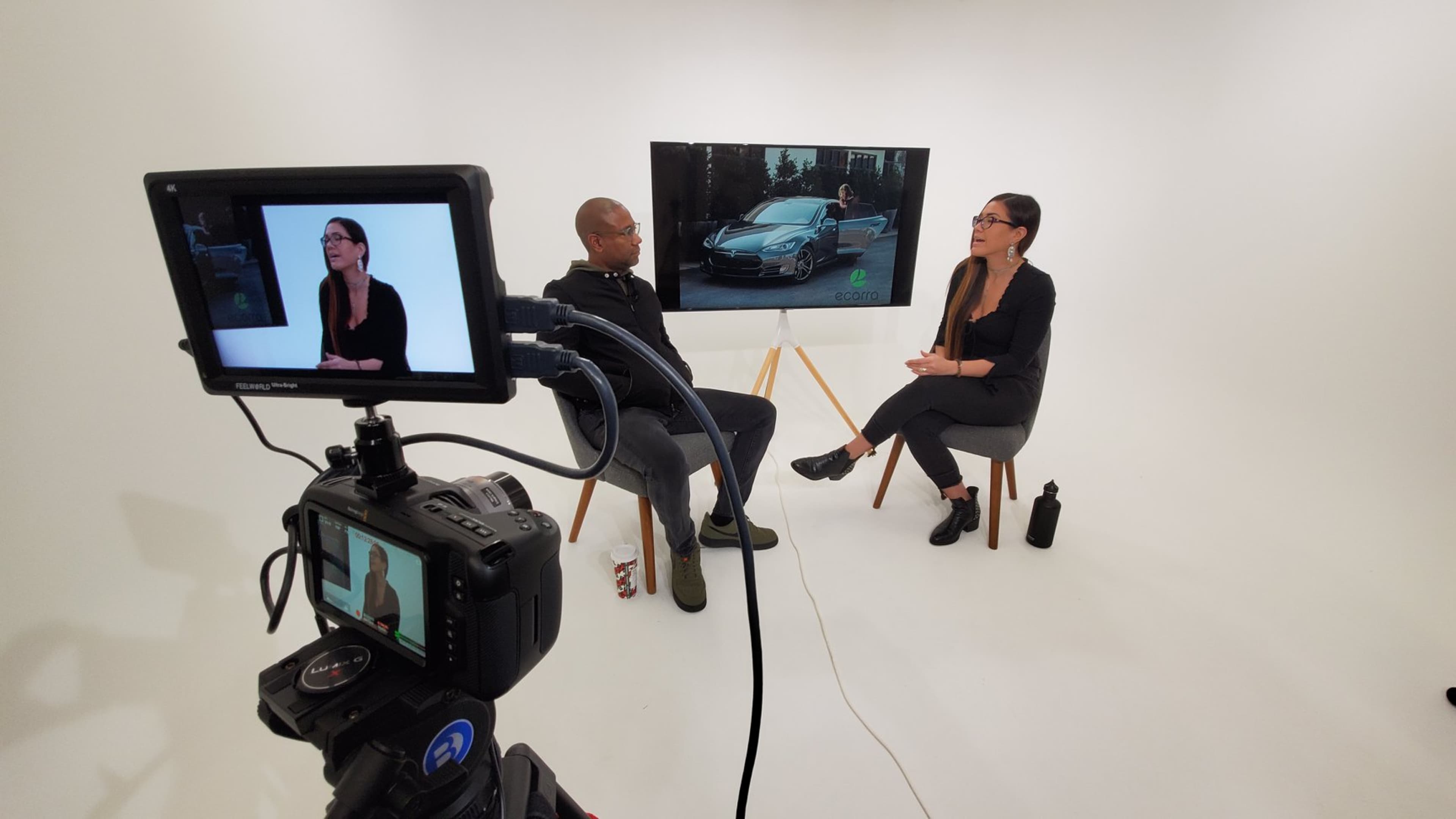 A man and a woman are seated in a brightly lit studio, discussing in front of a large screen displaying a car image.