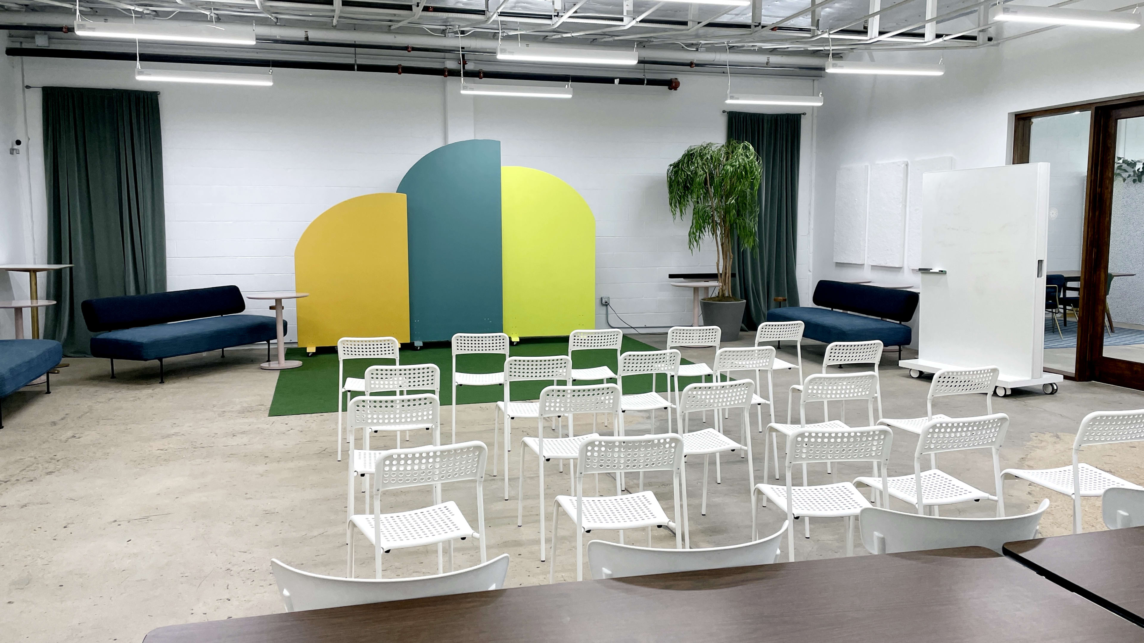 A modern meeting room features white chairs arranged in rows facing a colorful backdrop of yellow and green, with sofas and large windows in the background.
