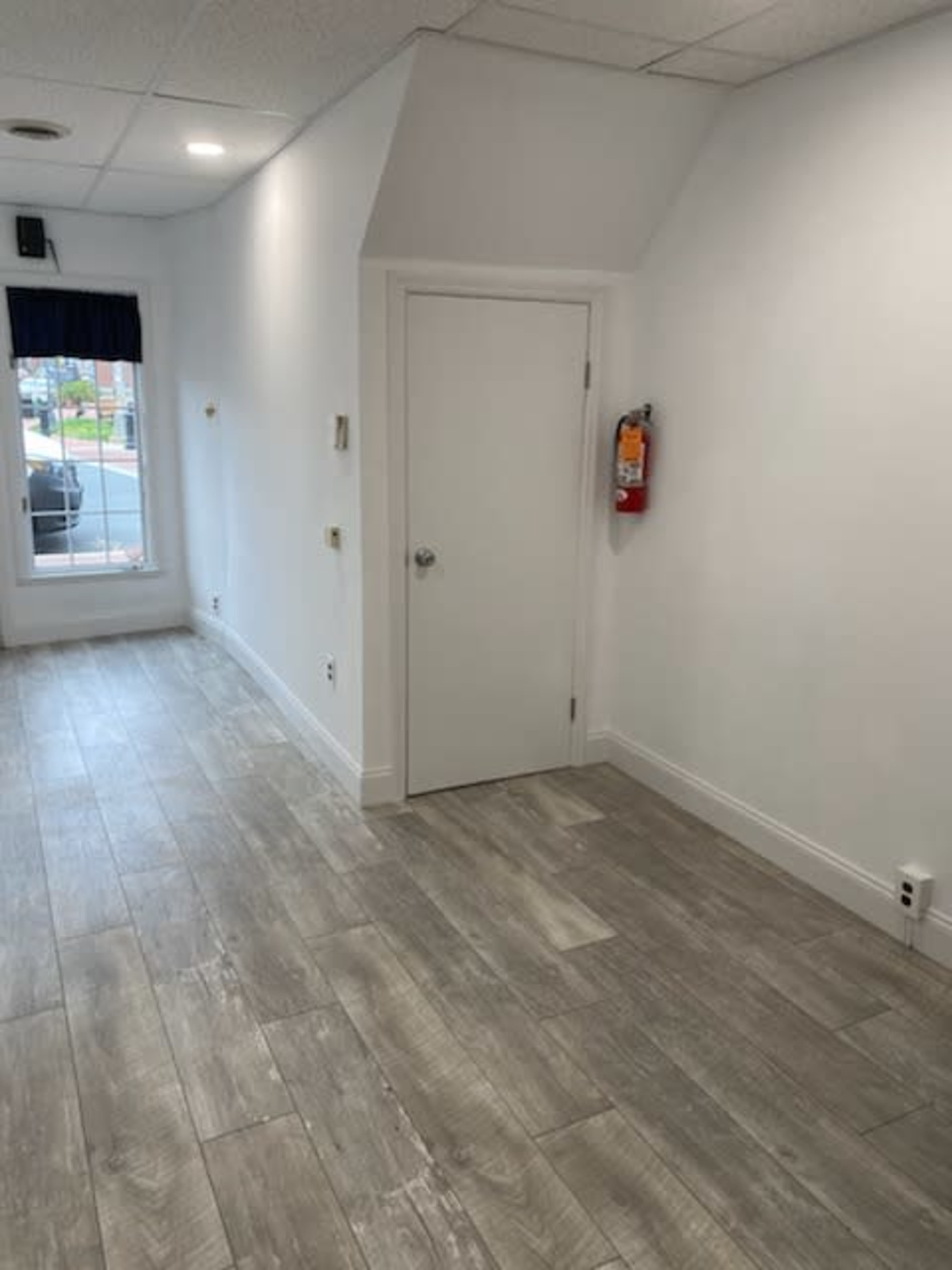 A vacant, well-lit room with light-colored walls, wooden flooring, and a closed door on one side.