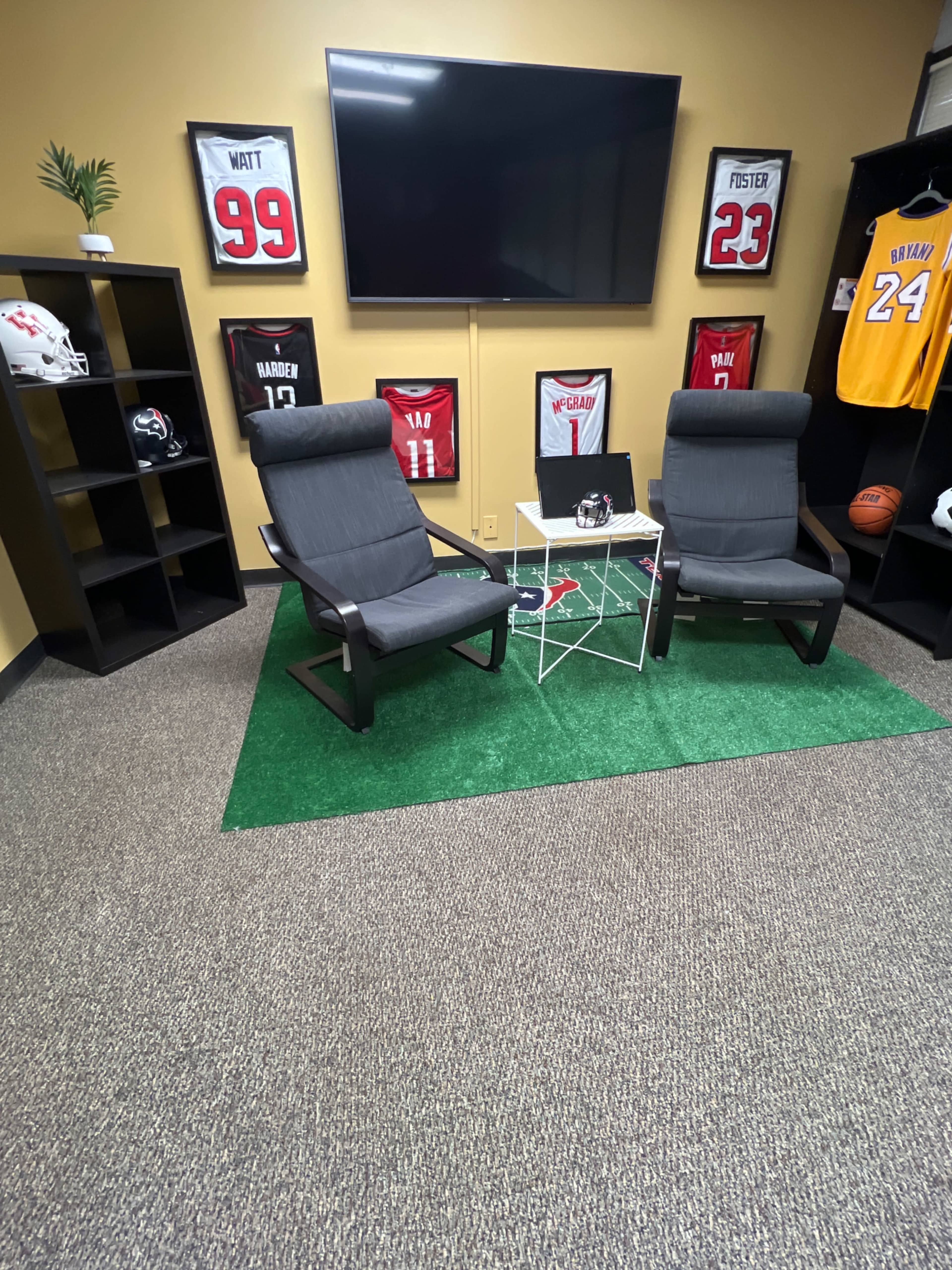 The scene features two gray chairs facing a small table on a green carpet, surrounded by framed jerseys on the walls and a large TV mounted above.