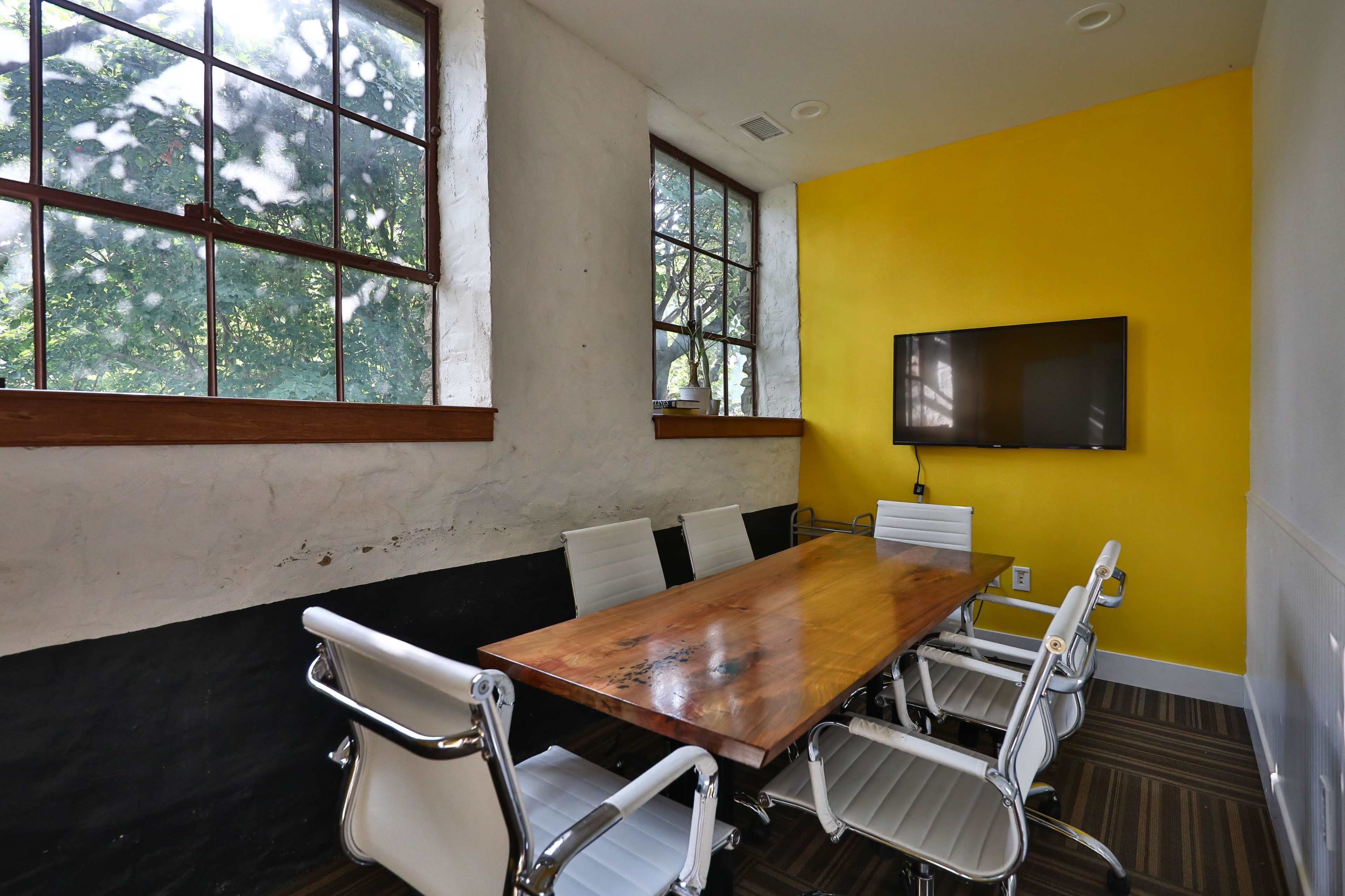 A small conference room features a wooden table surrounded by white chairs, set against a yellow wall and large windows.