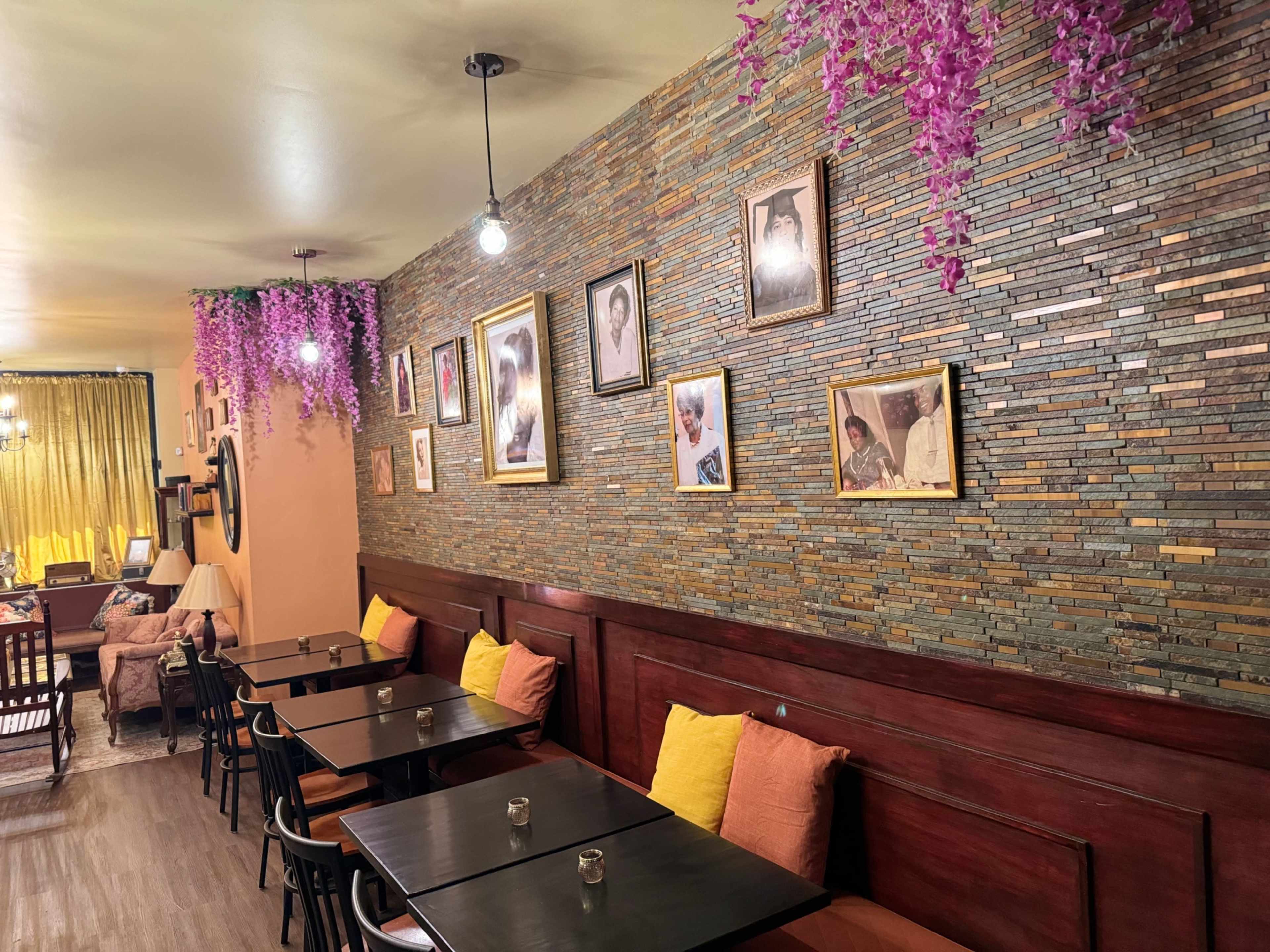 The image shows a café interior with a stone wall adorned with framed pictures and hanging purple flowers, while tables with black tops and colorful cushions line the seating area.