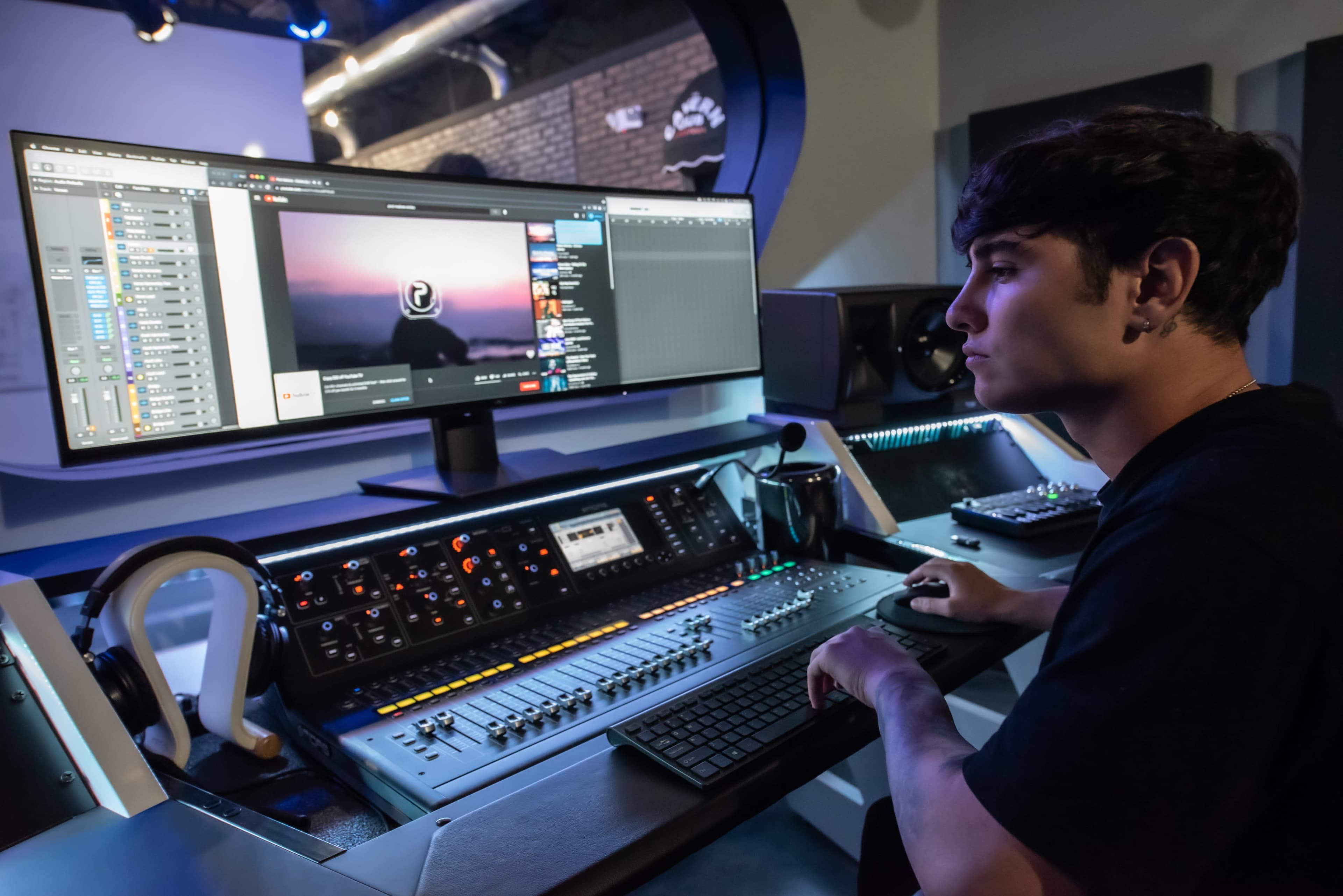 A young man sits at a modern audio production workstation, focused on a computer screen displaying music software.