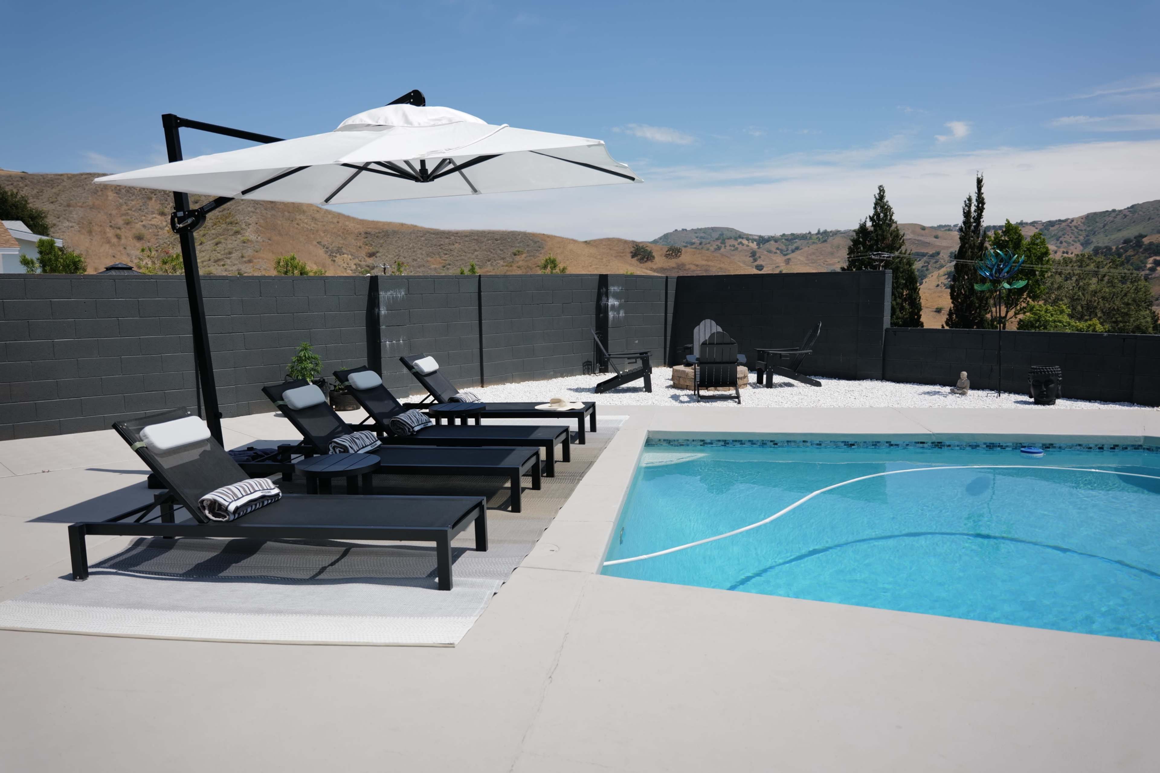 The image shows a swimming pool area with several lounge chairs, an umbrella, and a backdrop of hills under a clear blue sky.