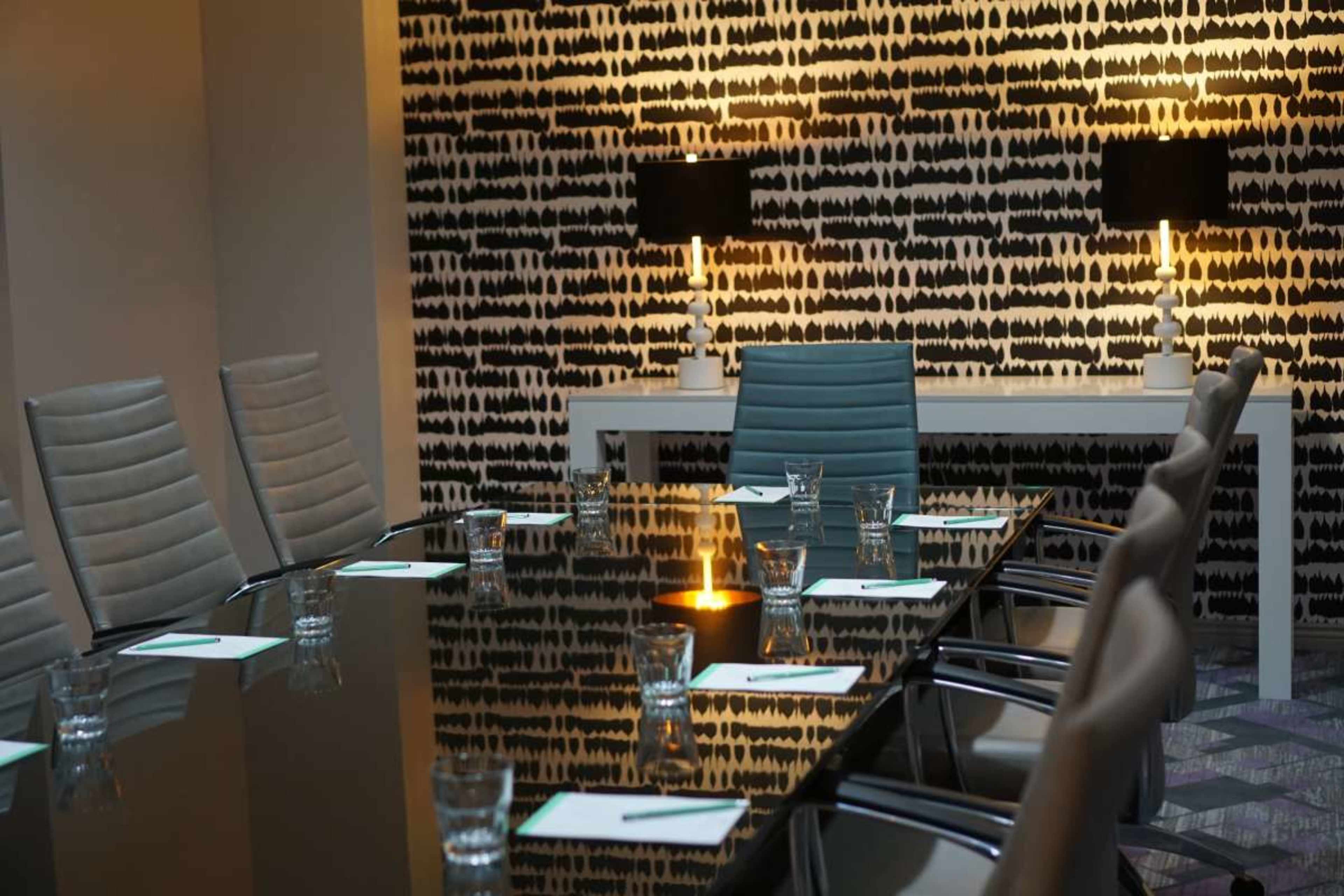 A modern conference room features a long glass table surrounded by sleek chairs, with a patterned wall and decorative lamps illuminating the space.
