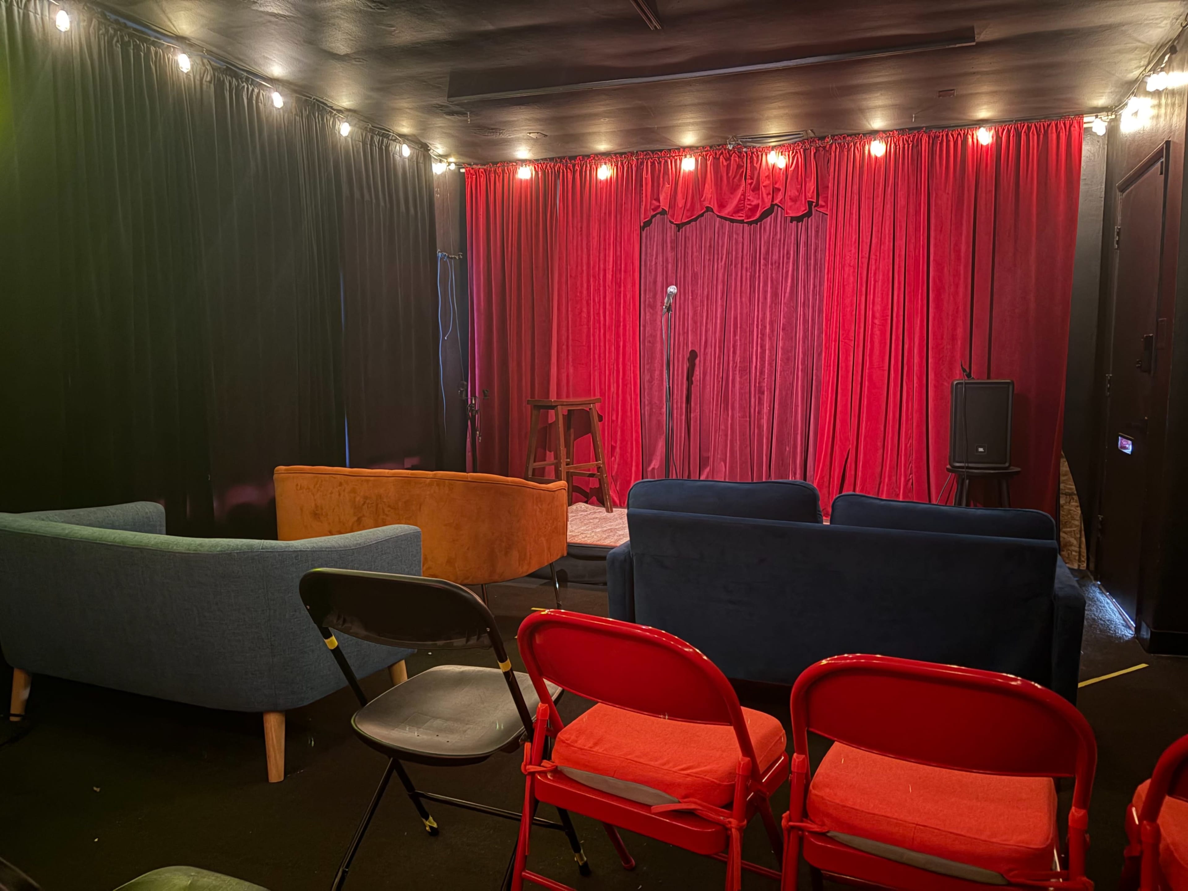 A small performance space features a stage with red curtains, a microphone stand, and a mix of colorful seating arranged in front.