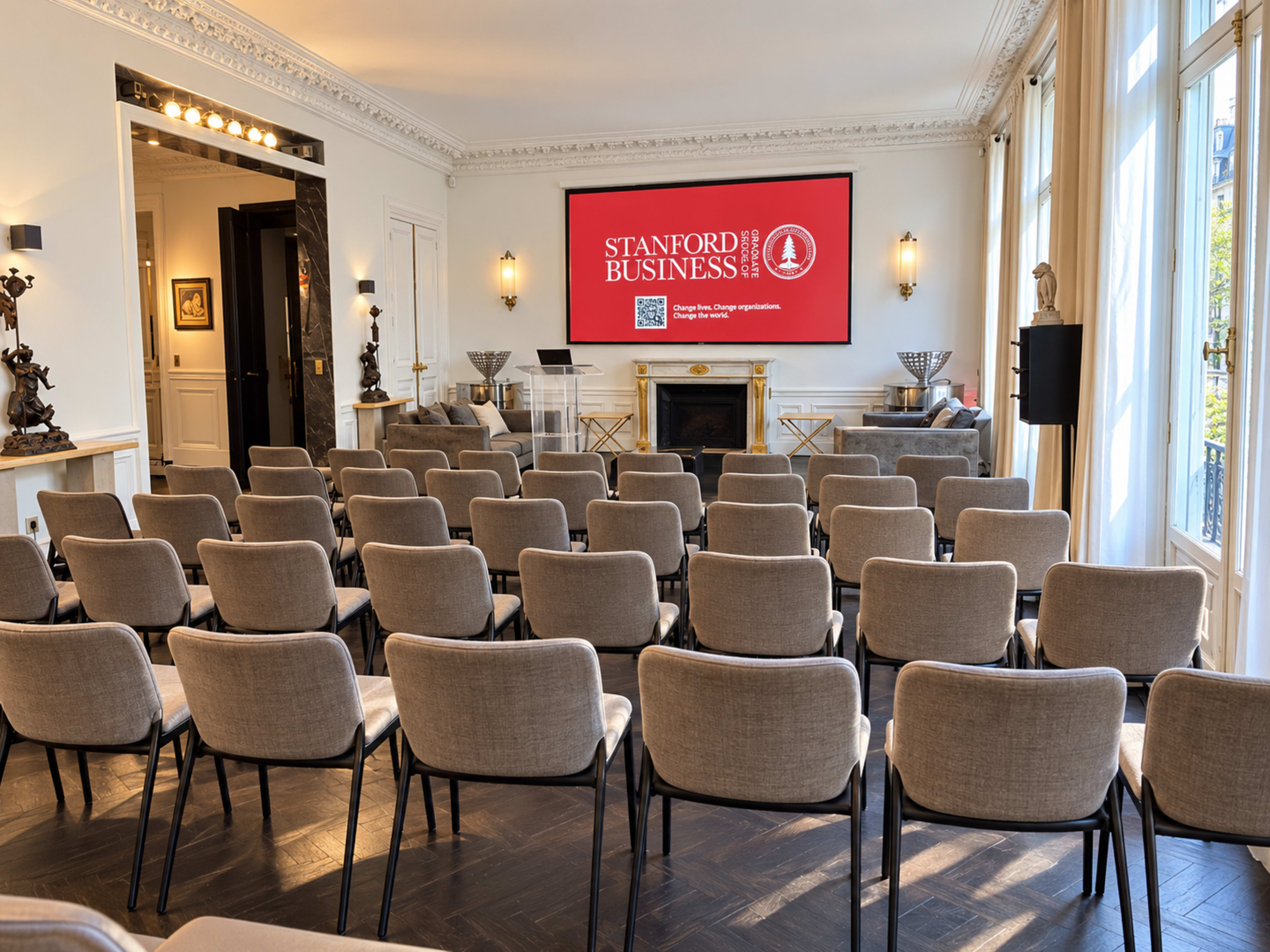 A conference room is set up with rows of beige chairs facing a large screen displaying the logo of Stanford Business, situated in a well-lit, elegantly decorated space.