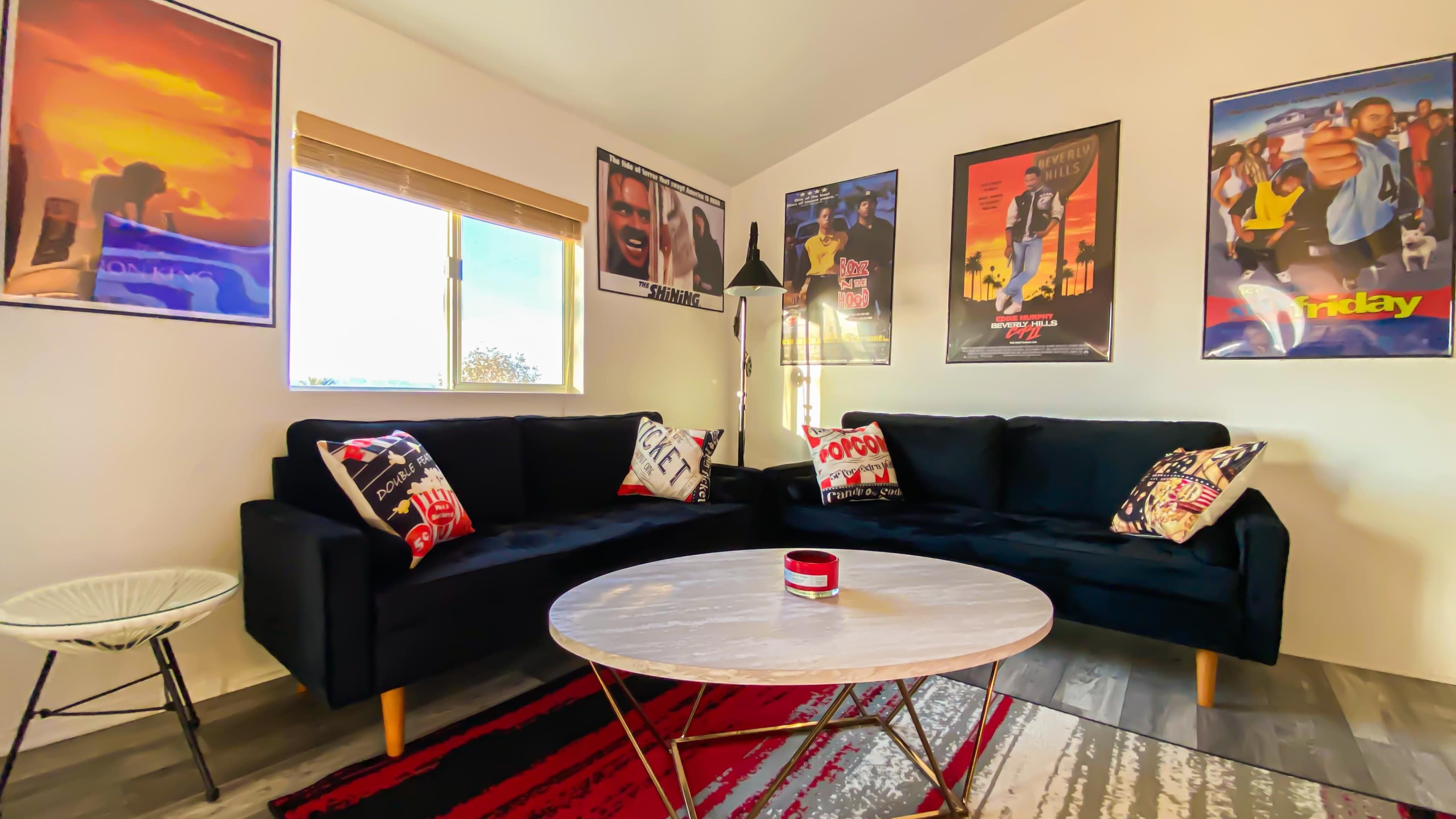 A cozy living room features two black sofas, a round marble coffee table, and decorated walls with various movie posters.