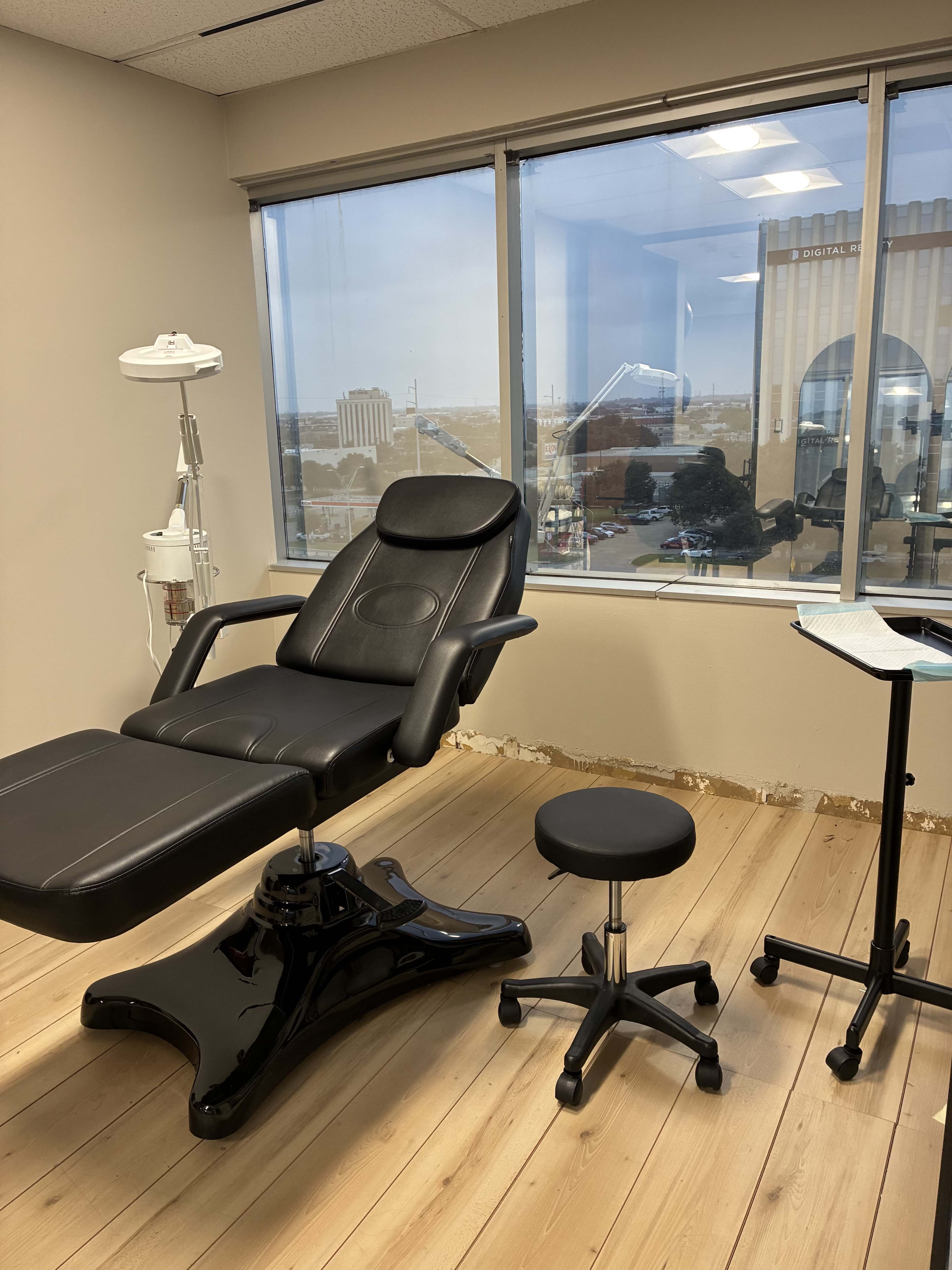 The image shows a cosmetic treatment room with a reclining chair, a small stool, and a treatment cart beside a large window.