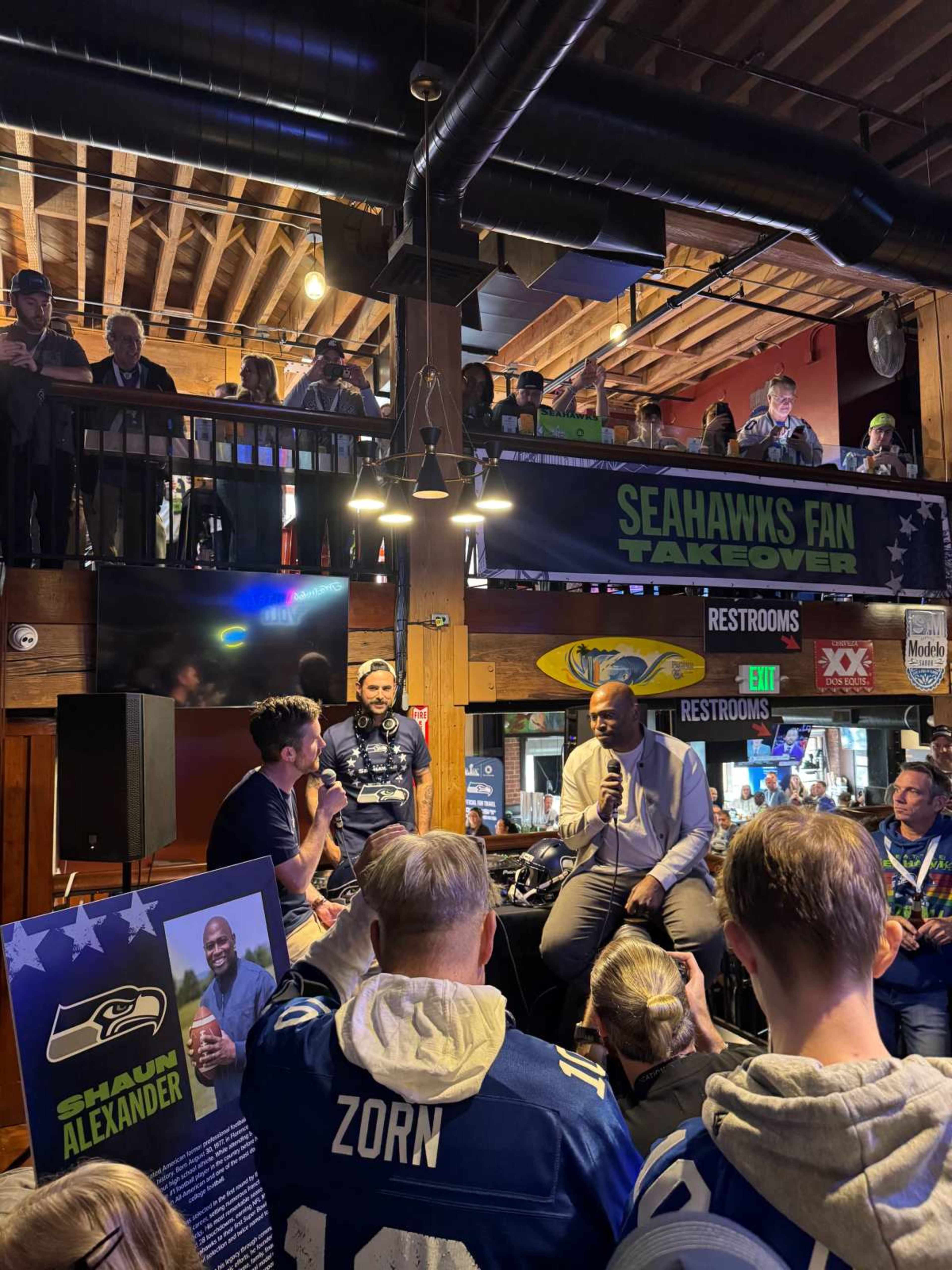 A crowd gathers in a venue, as two men sit on a stage discussing Seahawks player Shaun Alexander, while fans in jerseys watch from the balcony.