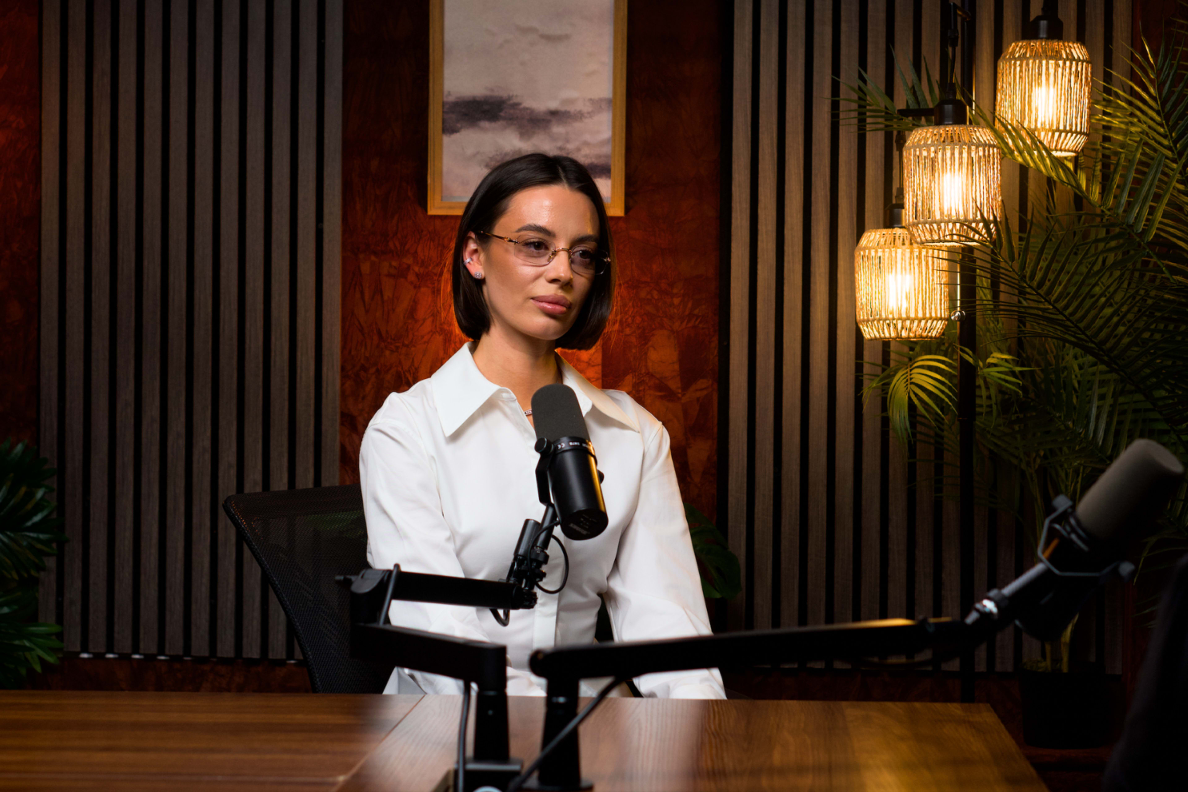 A woman wearing a white shirt sits at a desk with microphones in a well-lit studio featuring wood paneling and hanging lights.