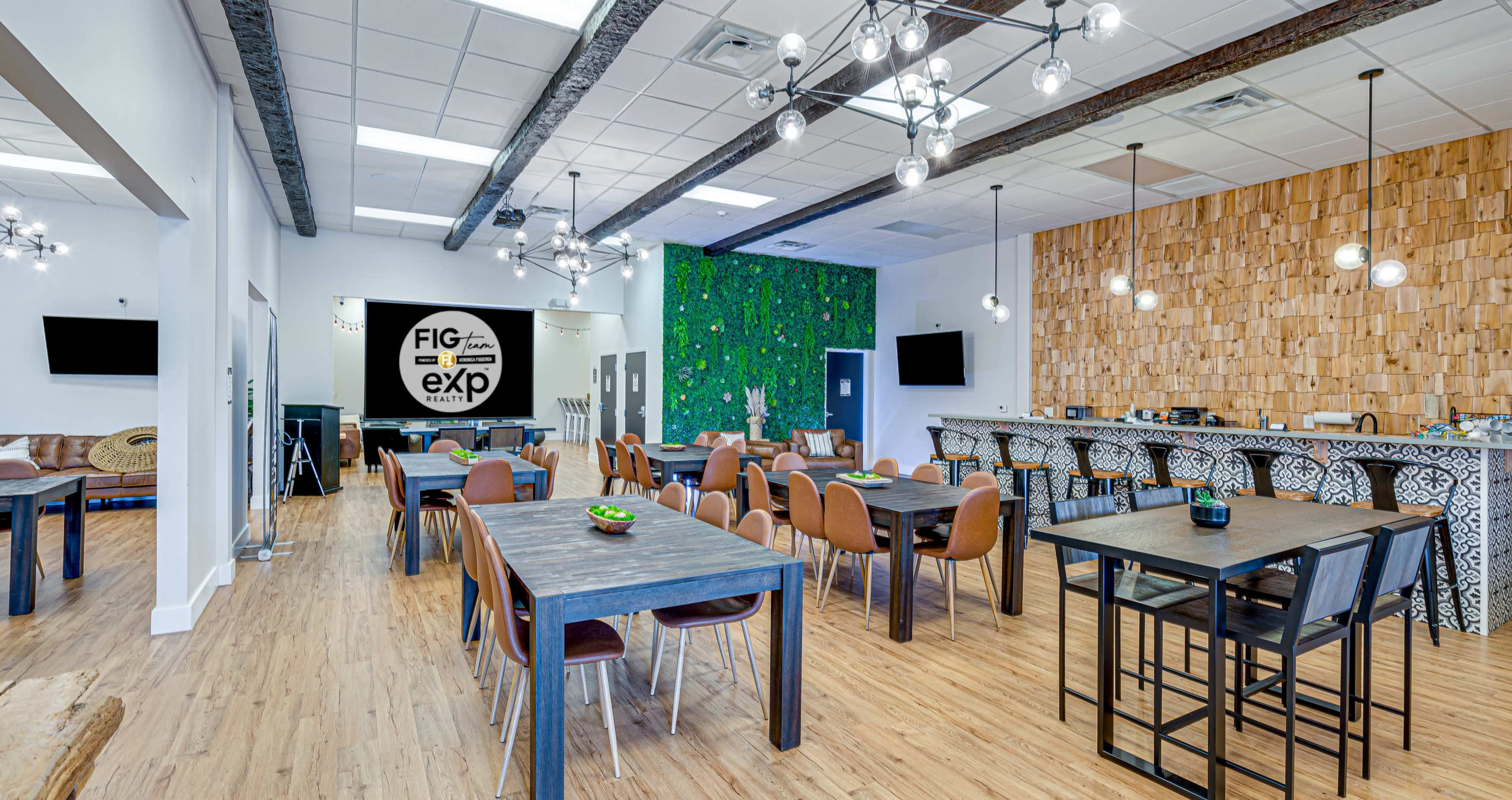 A modern co-working space with wooden floors, multiple tables arranged for dining or working, plants on the walls, and a bar area with decorative pendant lighting.
