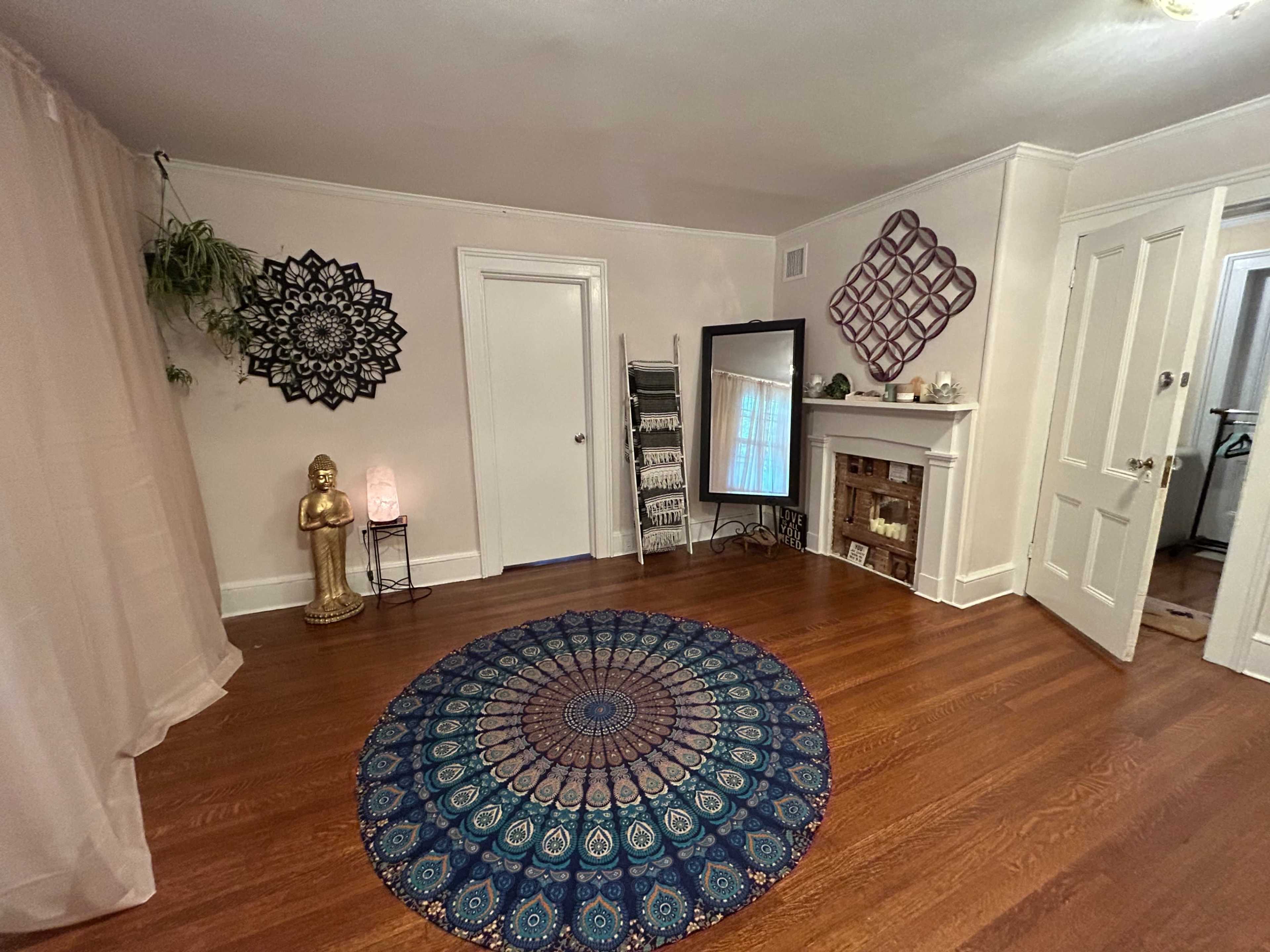 The room features a wooden floor, centered with a circular blue and teal patterned rug, a mirror, a decorative statue, and various wall decorations.