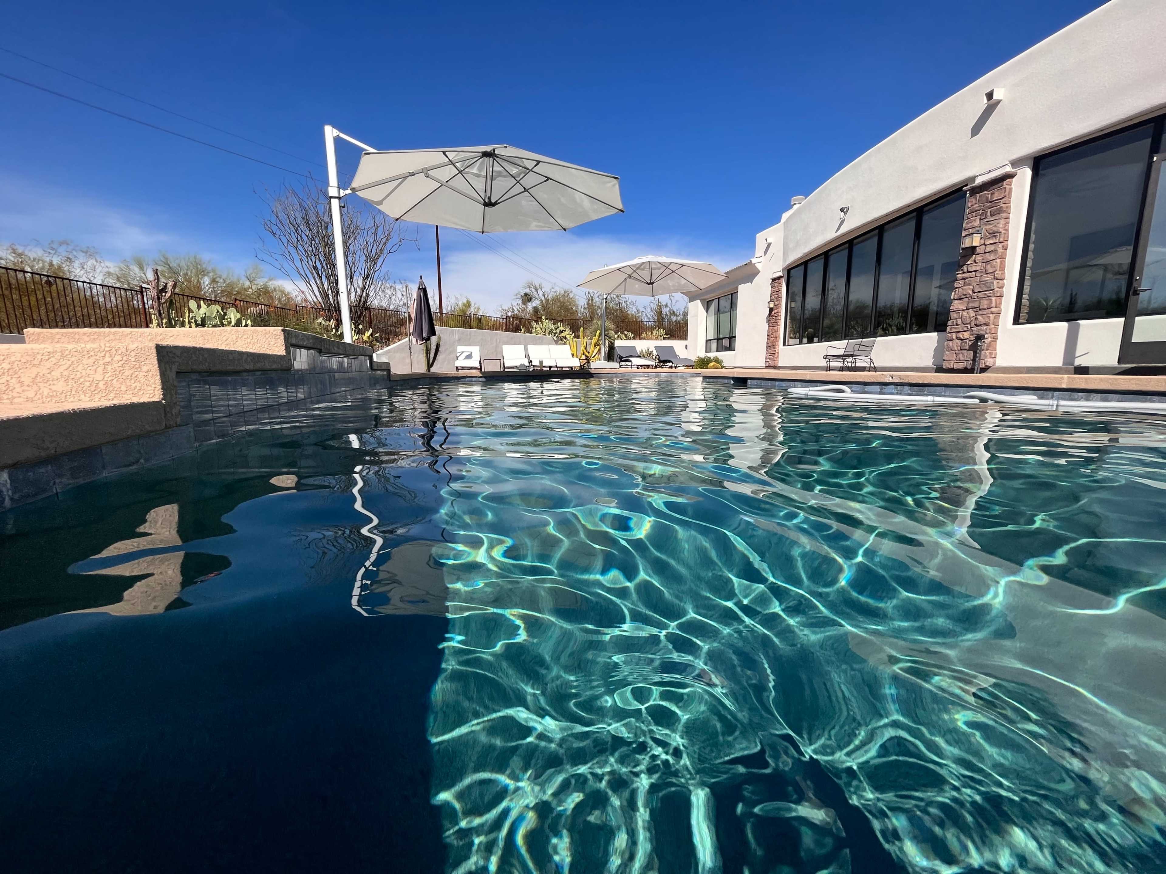 The image shows a clear swimming pool with reflections of umbrellas and a house against a blue sky.