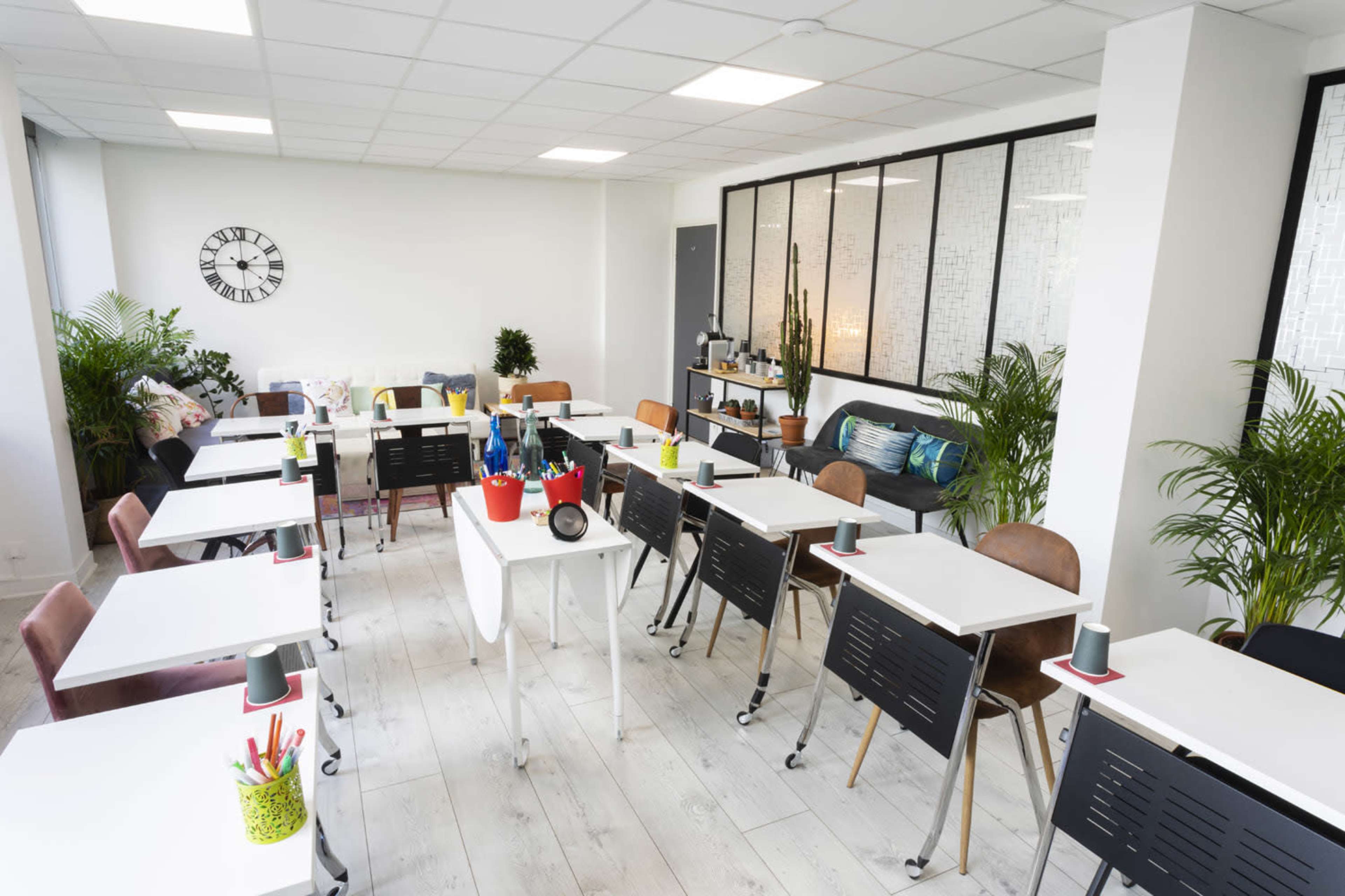 A modern classroom setup with multiple tables and chairs arranged neatly, surrounded by plants and featuring a wall with large windows.
