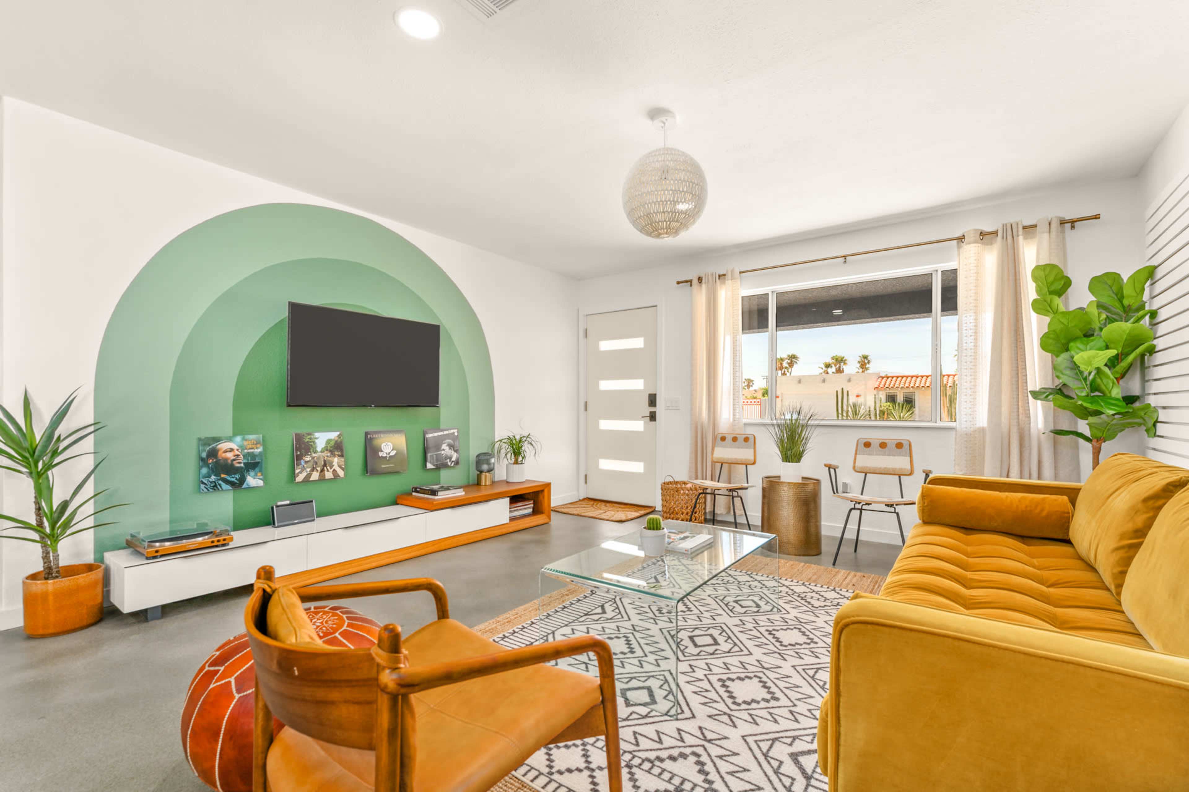 A modern living room features a green arc wall design, a television mounted above a low white console, a glass coffee table, and stylish seating arrangements, complemented by large windows and indoor plants.
