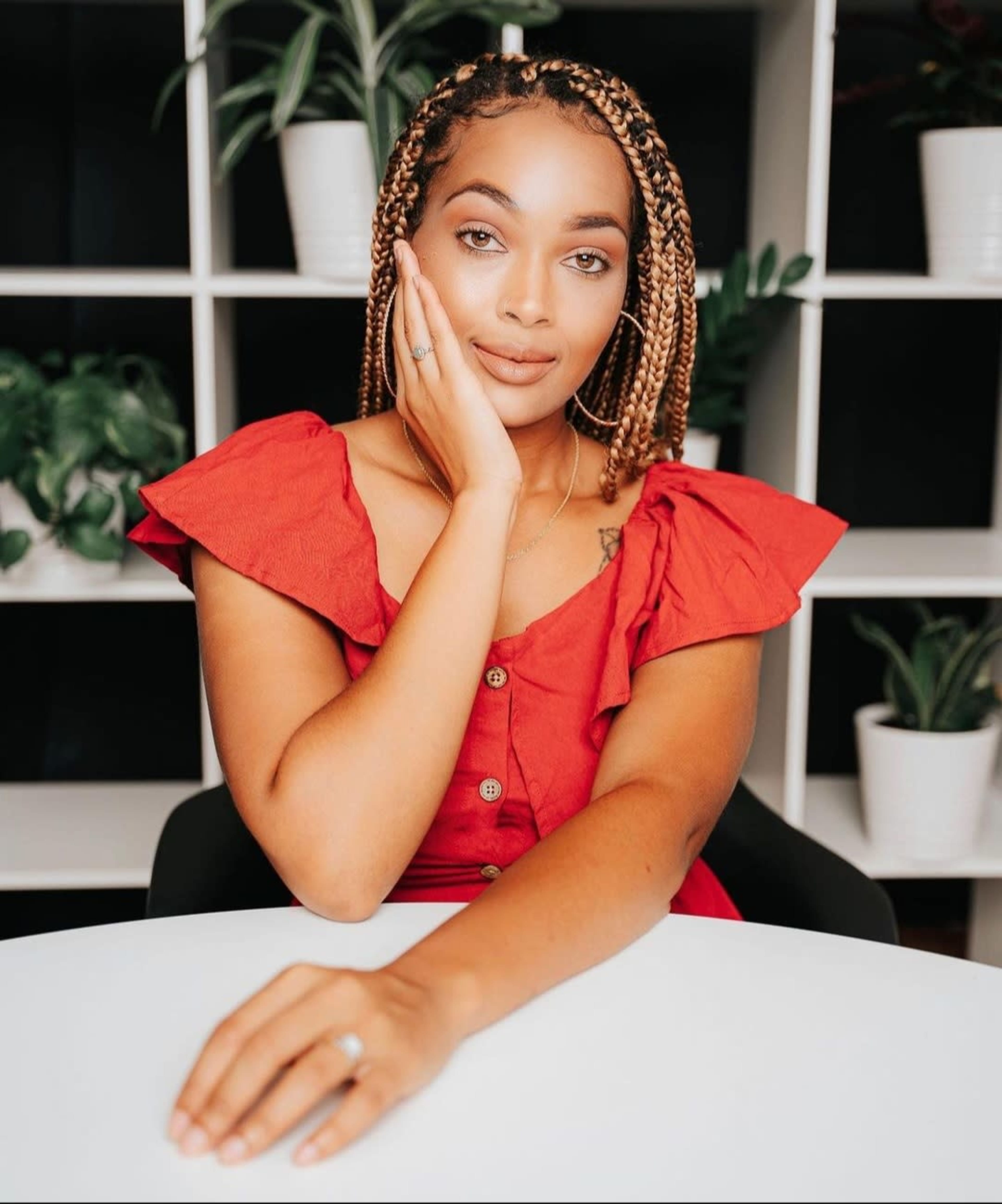 A woman with braided hair sits at a table, resting her chin on her hand, in front of a backdrop of potted plants and shelves.