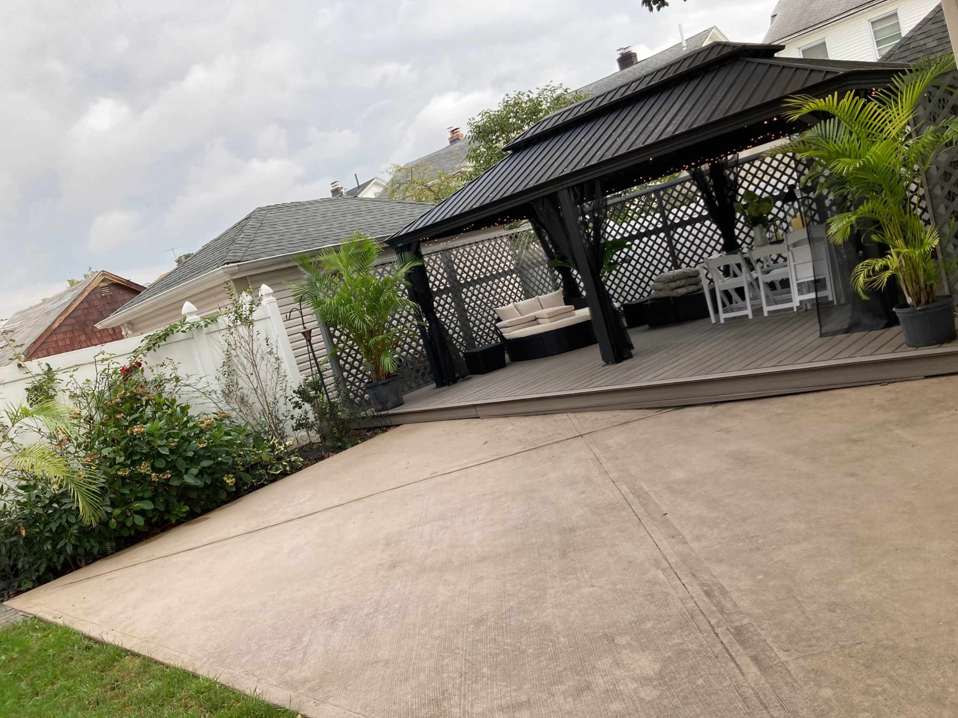 A covered outdoor gazebo with a wooden deck is set against a backdrop of residential homes and greenery.