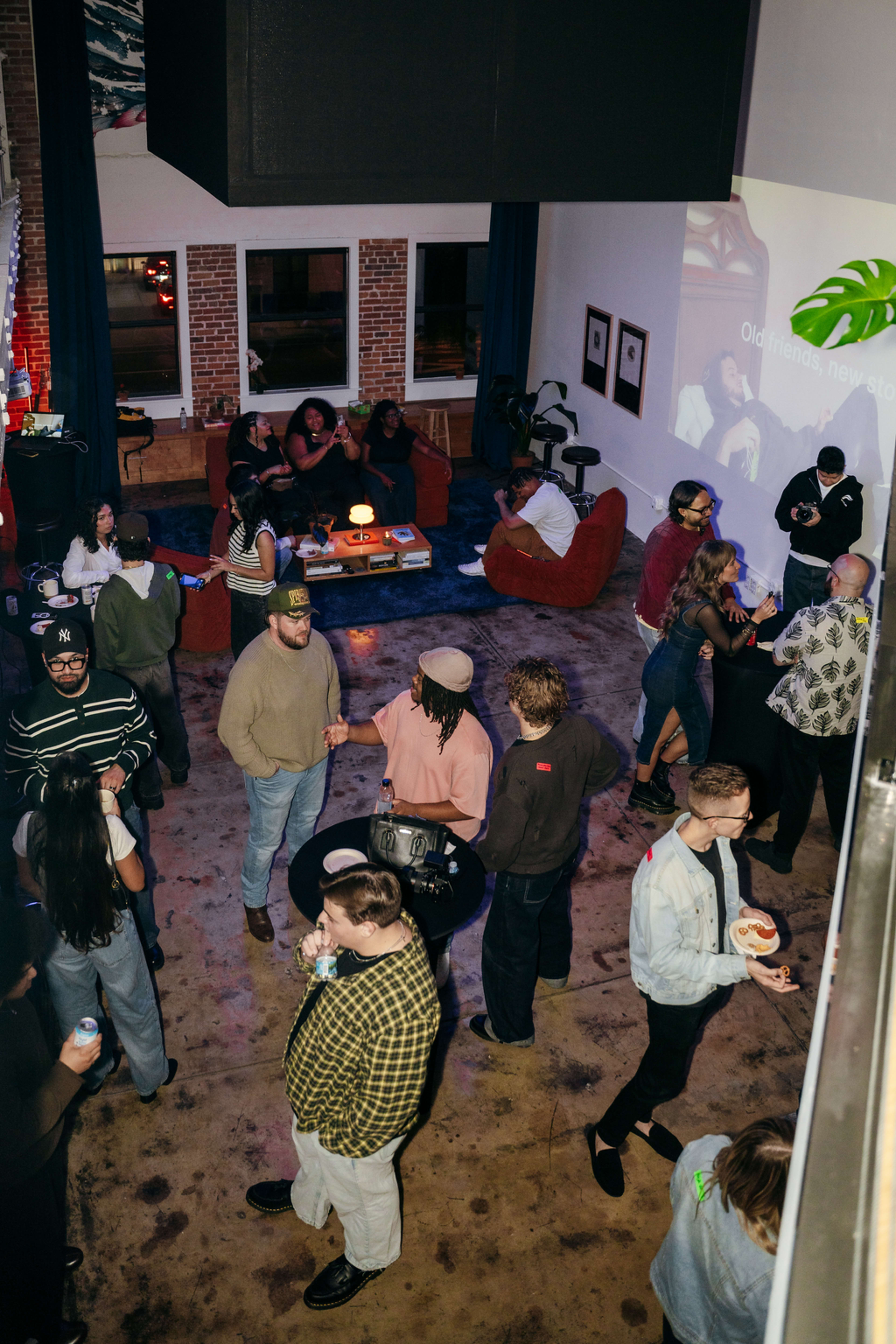 A crowded indoor gathering features people socializing, seated on sofas, and watching a projection in a stylish venue with exposed brick walls.