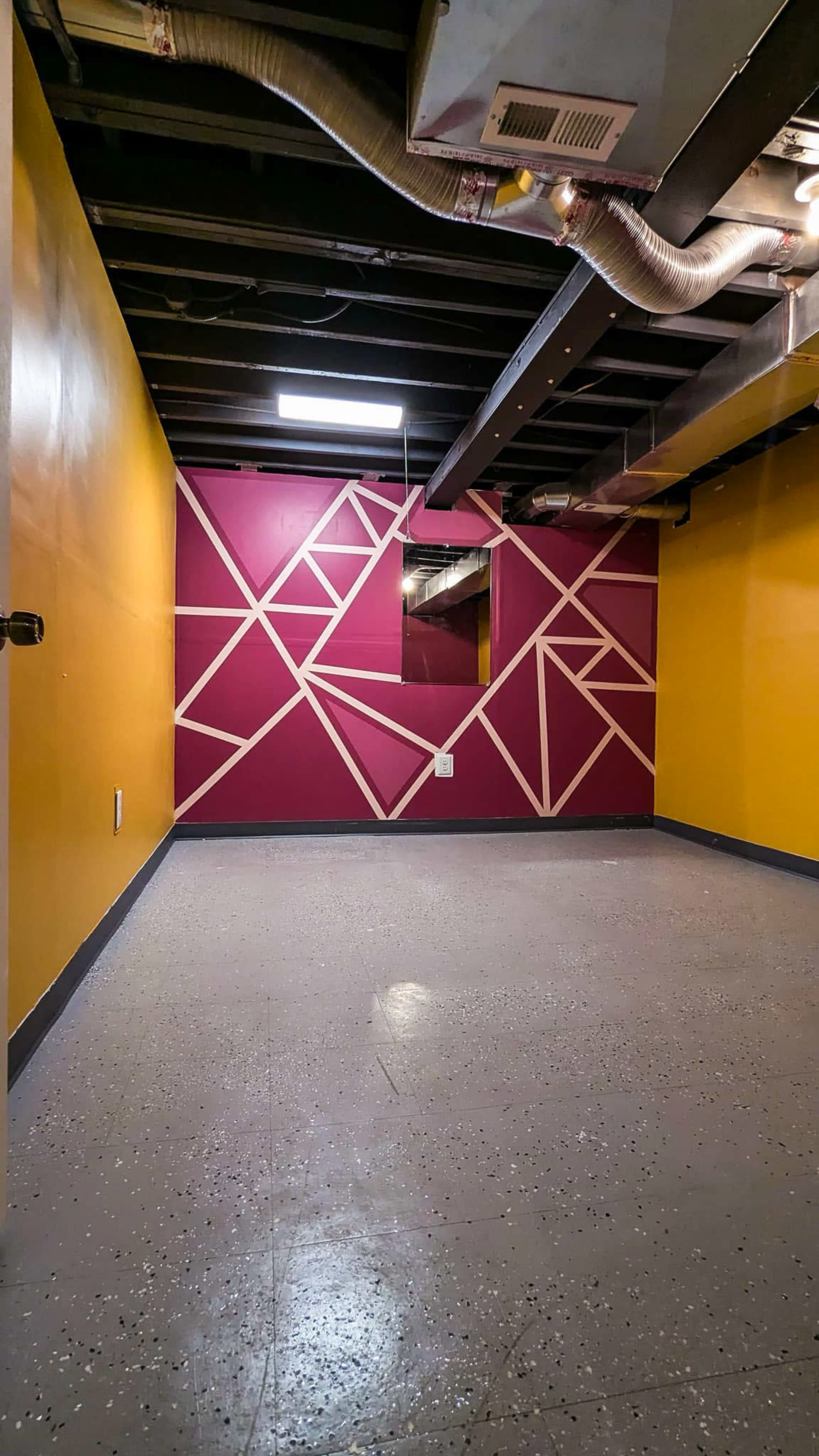 The image shows an empty basement room with yellow and maroon walls featuring a geometric design, and a small mirror attached to one of the walls.