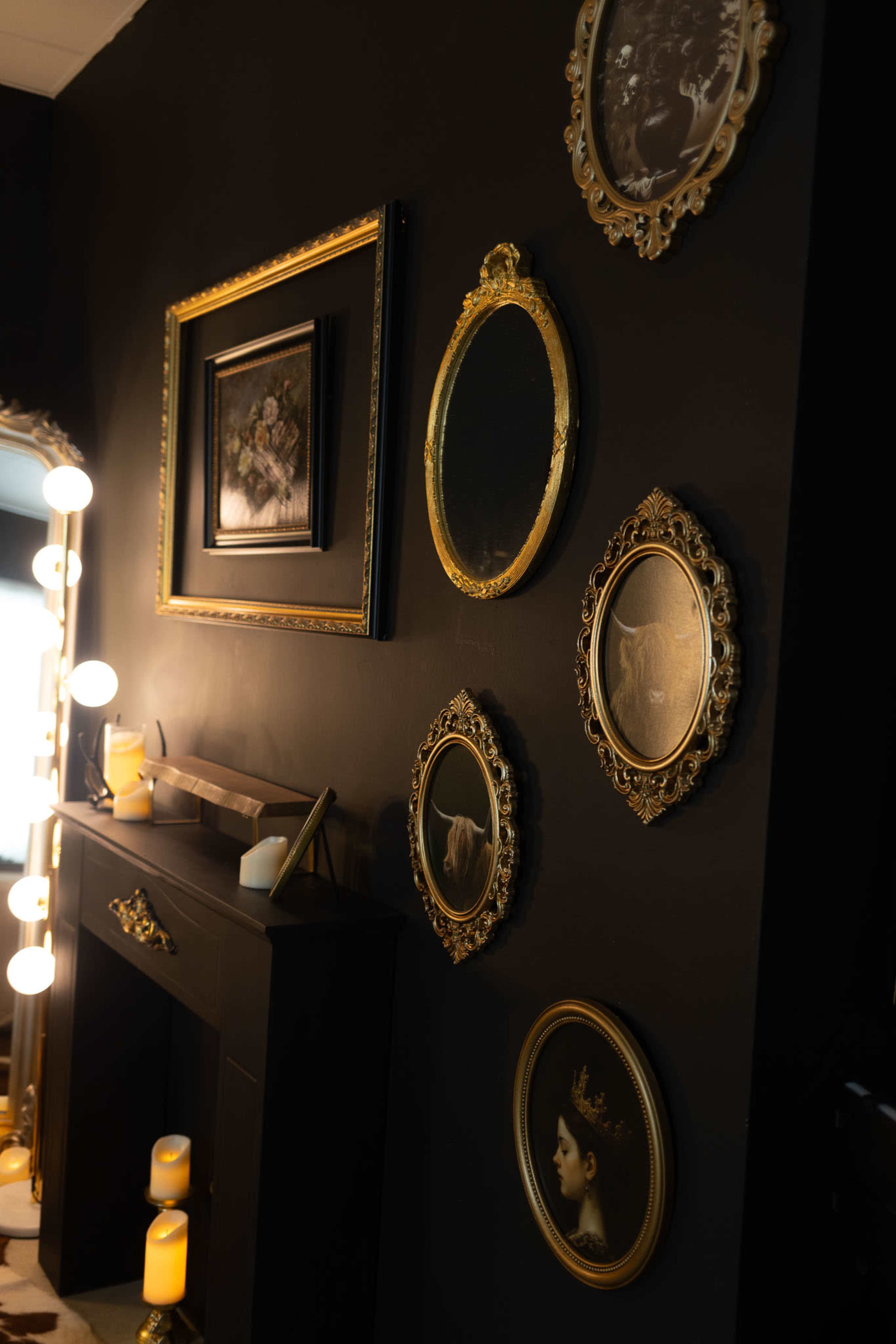 The image features a dark wall adorned with several ornate, round picture frames displaying various artworks, alongside a decorative mirror and candles on a mantelpiece.
