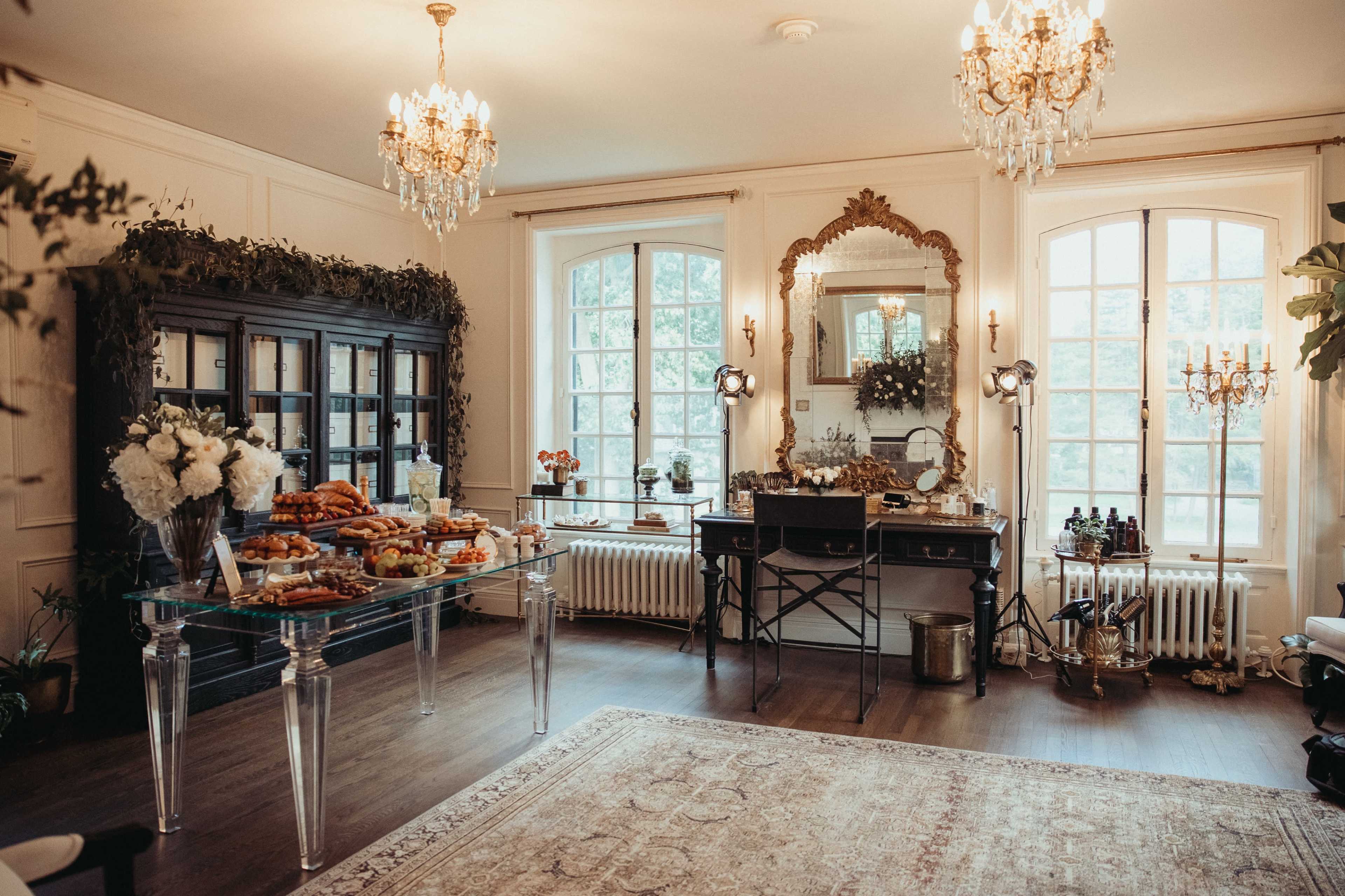 The image shows a elegantly decorated room featuring a table with assorted pastries, a mirror, and large windows allowing natural light, complemented by decorative chandeliers and greenery.