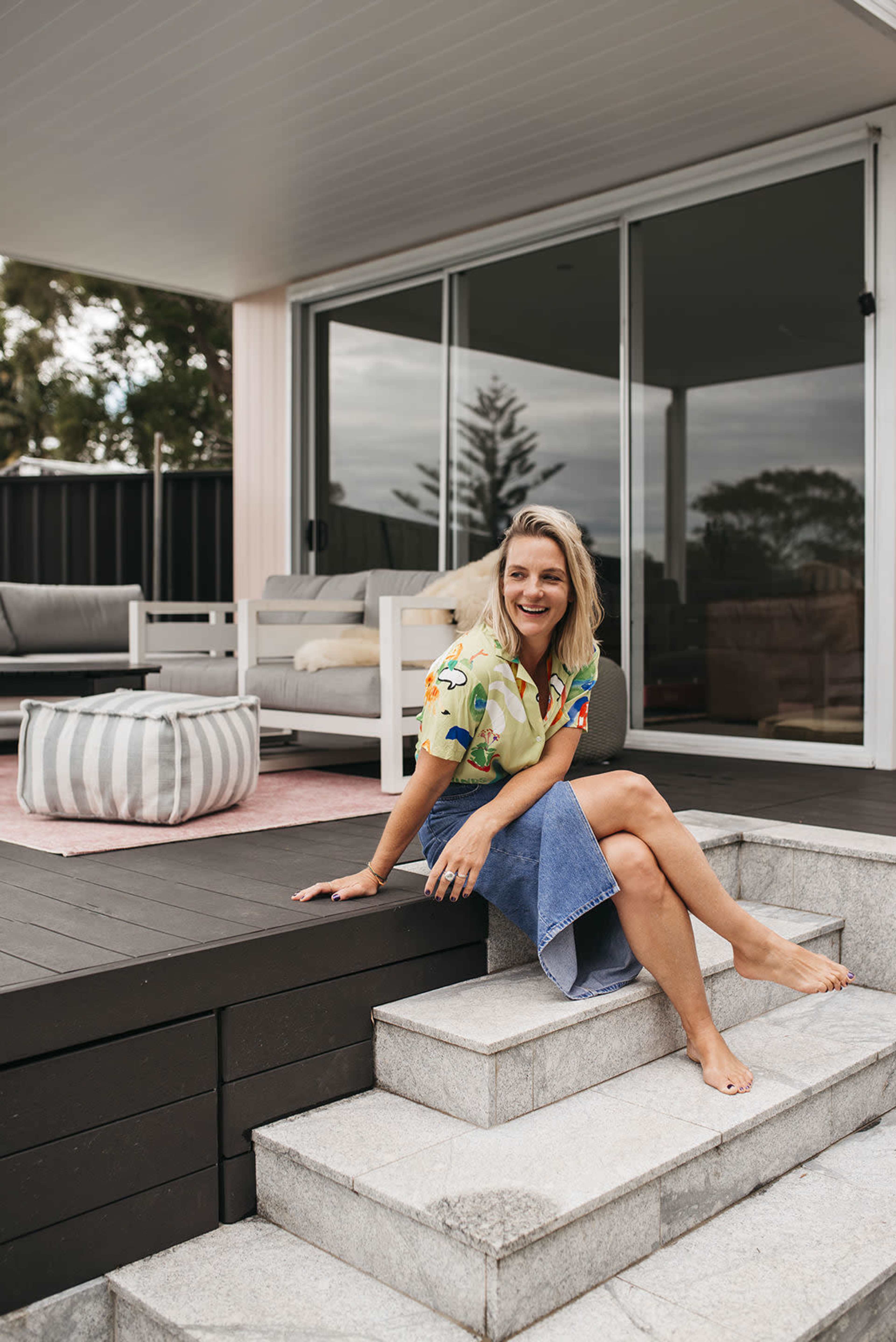 A woman sits on the steps of a patio, smiling and dressed in a colorful top and denim skirt, with a modern outdoor setting visible behind her.