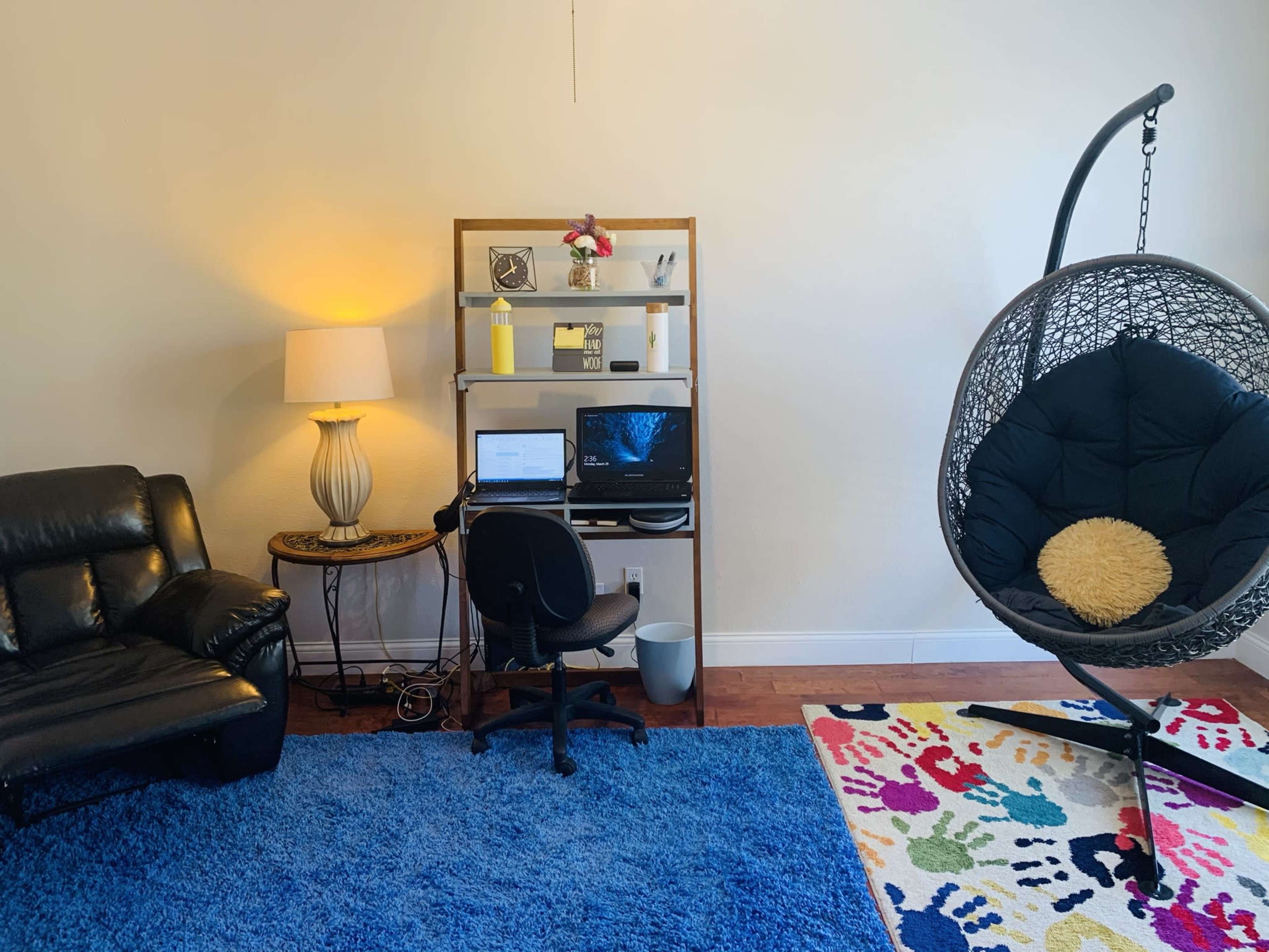 A living space featuring a black leather couch, a desk with a computer, and a hanging chair, all on a vibrant blue rug.