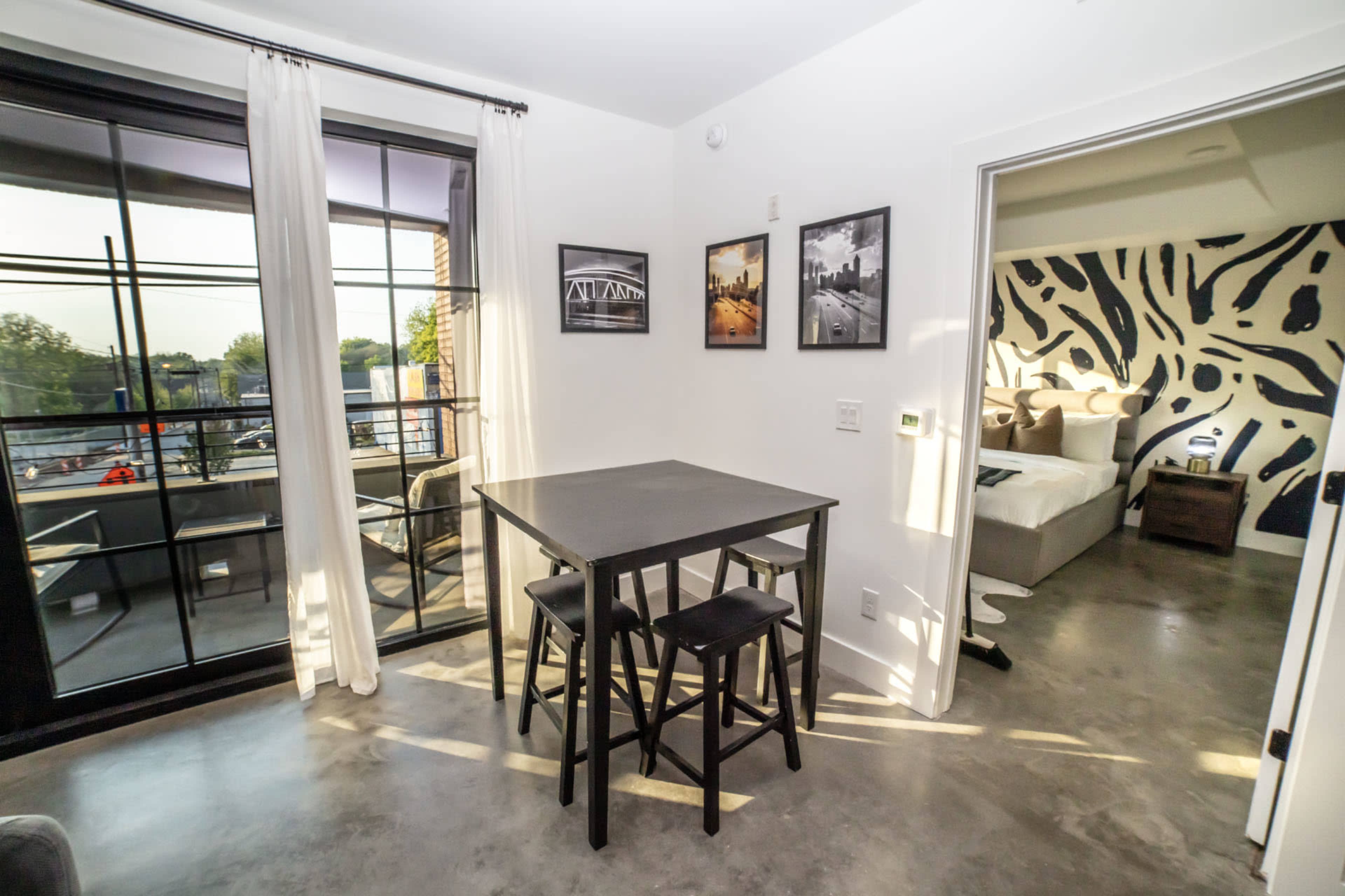 The image shows a modern apartment interior featuring a small dining table with stools, large windows, and a bedroom visible through an open doorway.