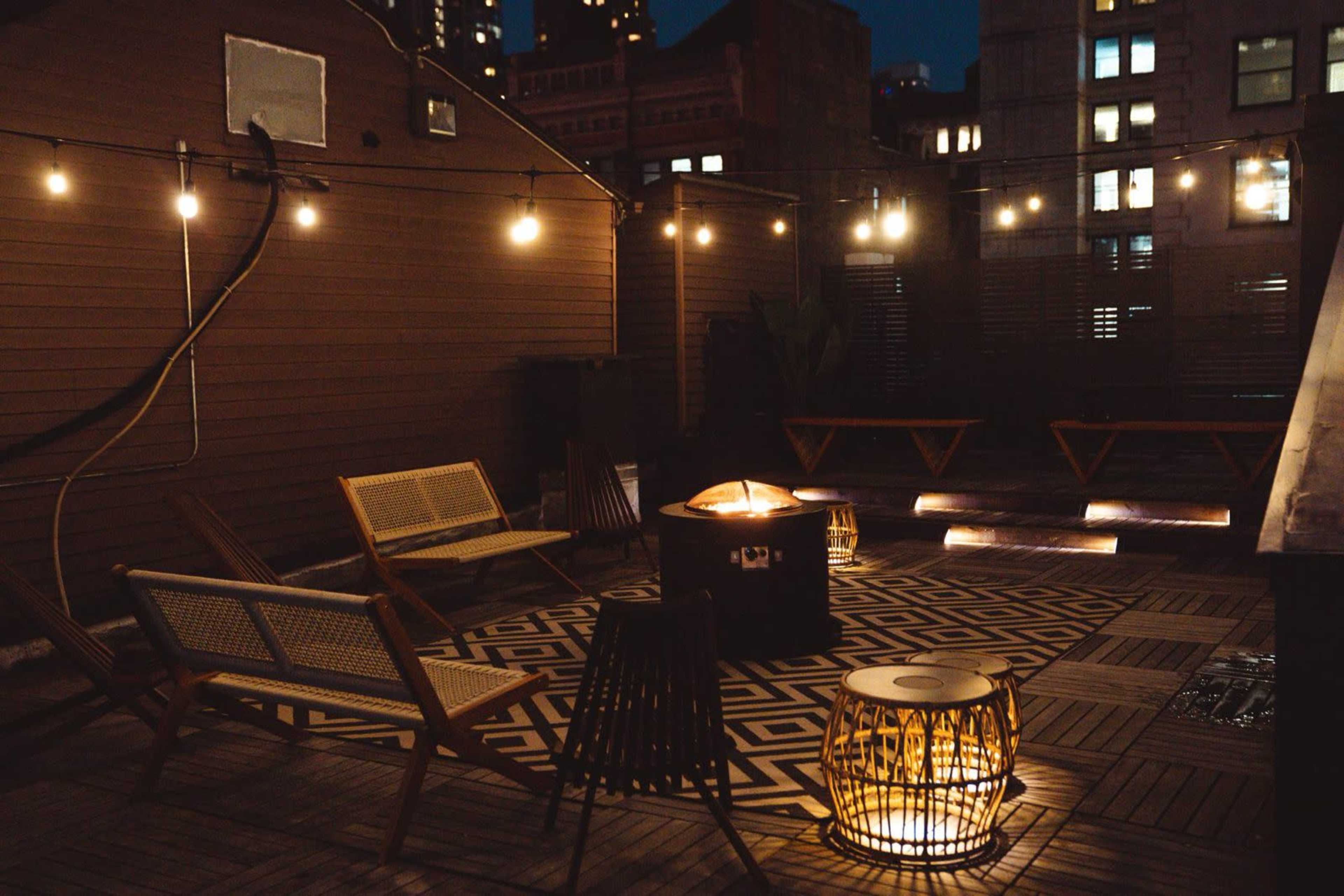 The image shows a rooftop space at night with string lights, patterned flooring, and various seating options around a fire pit.