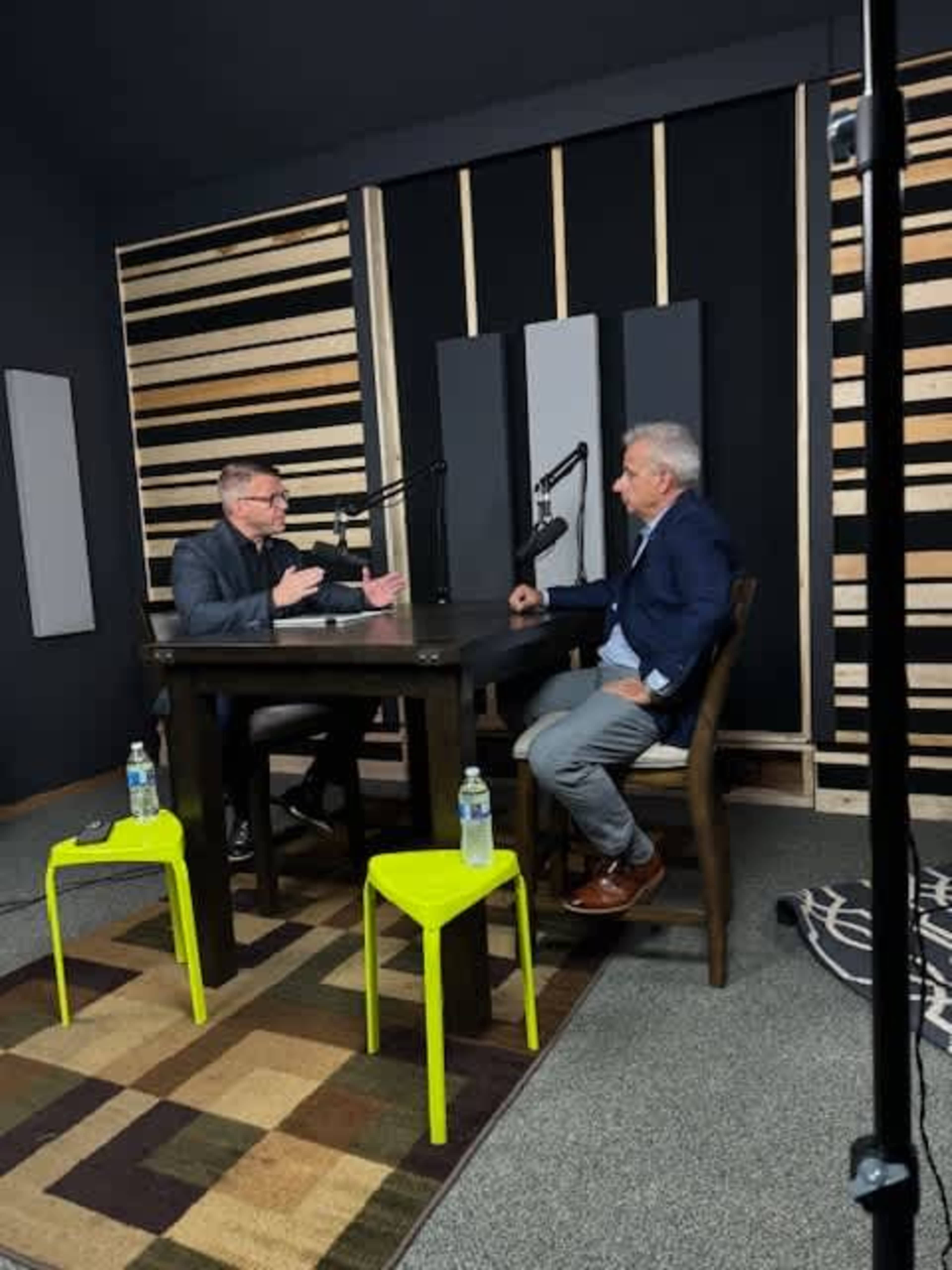 Two men are seated at a table in a podcast studio, engaged in conversation, while microphones are positioned in front of them.