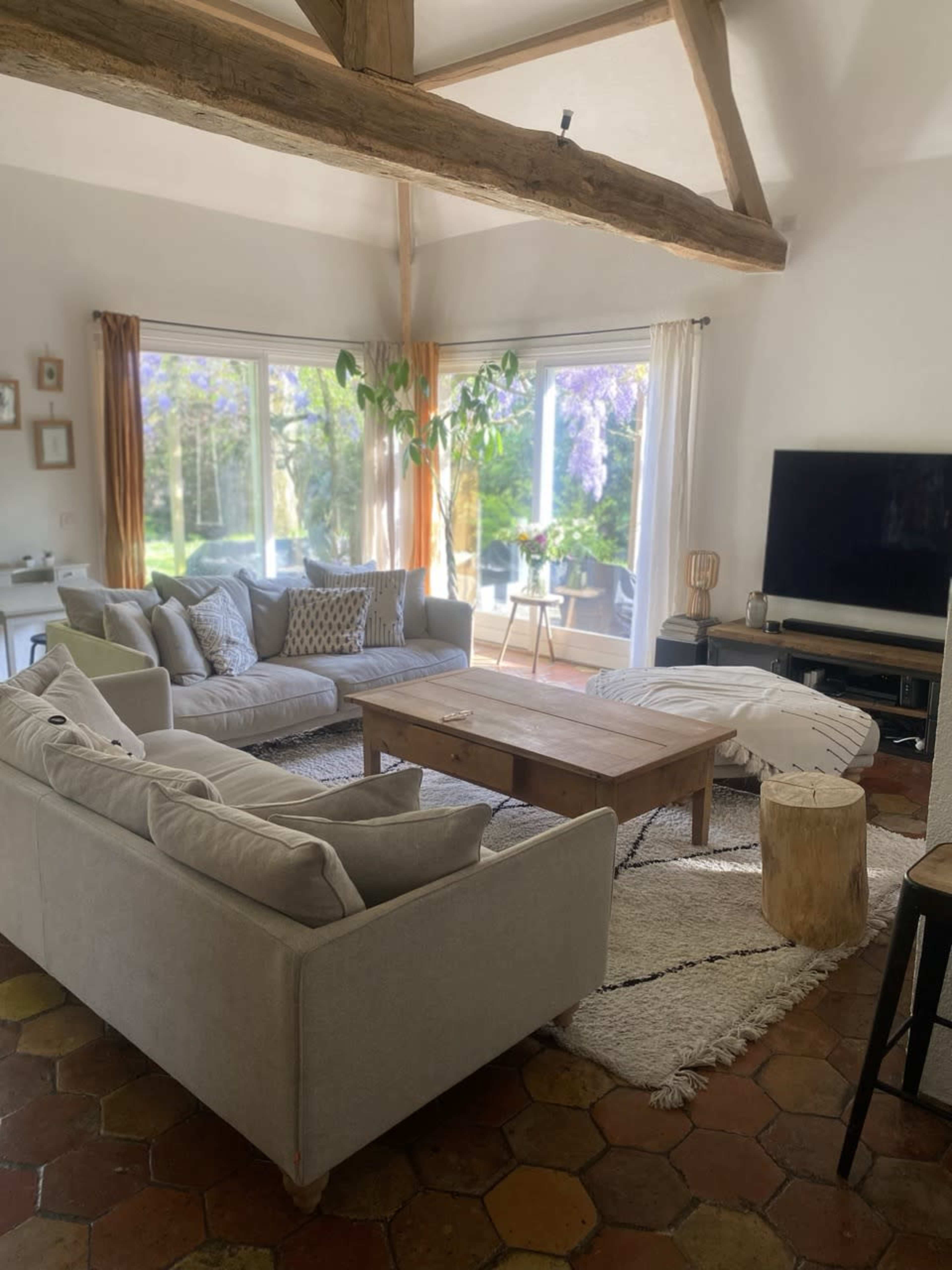 A cozy living room with two light-colored sofas arranged around a wooden coffee table, featuring a large window that opens to a green outdoor space.