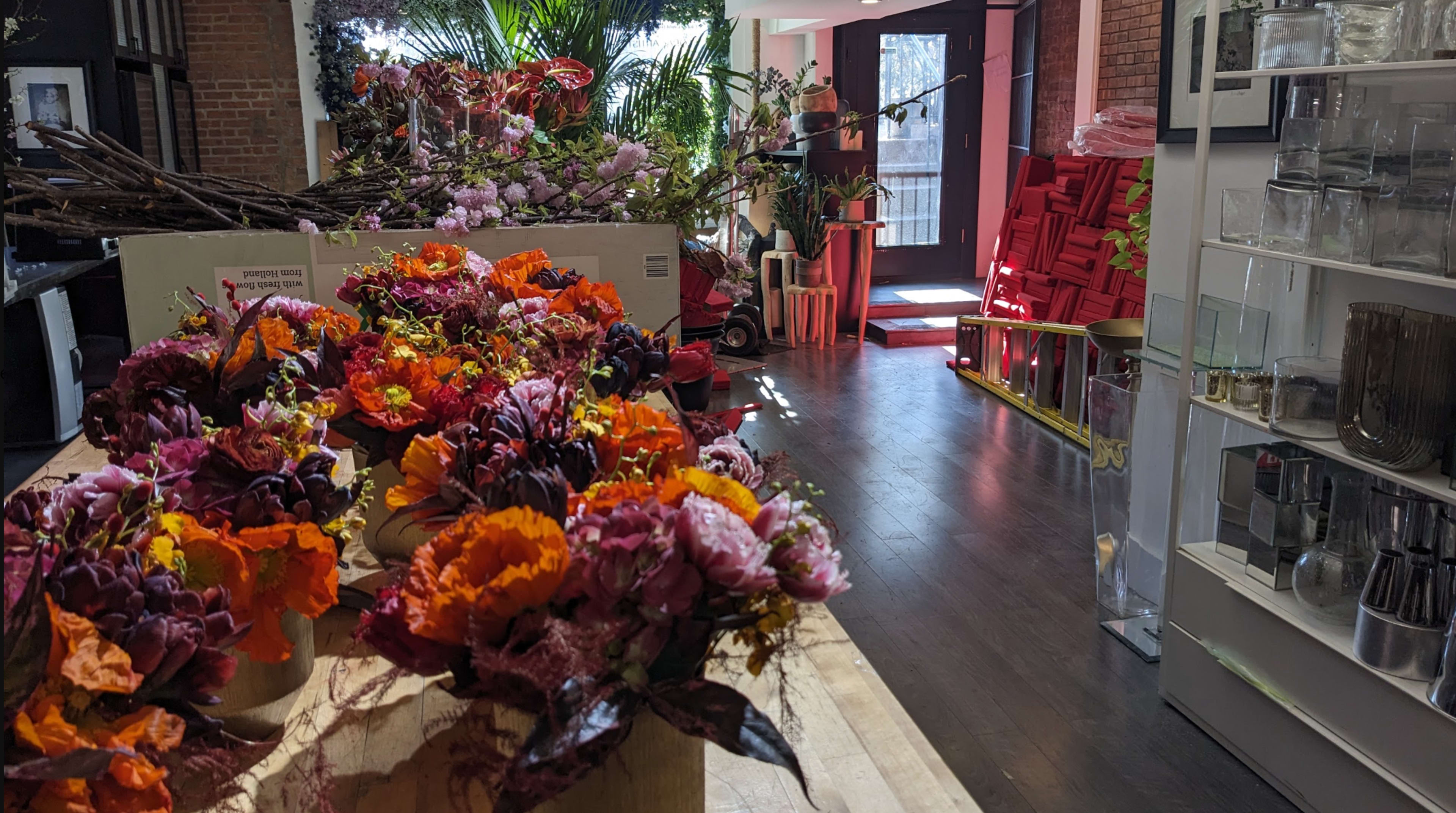The image shows a flower shop interior with vibrant floral arrangements in the foreground and red decorative items in the background.