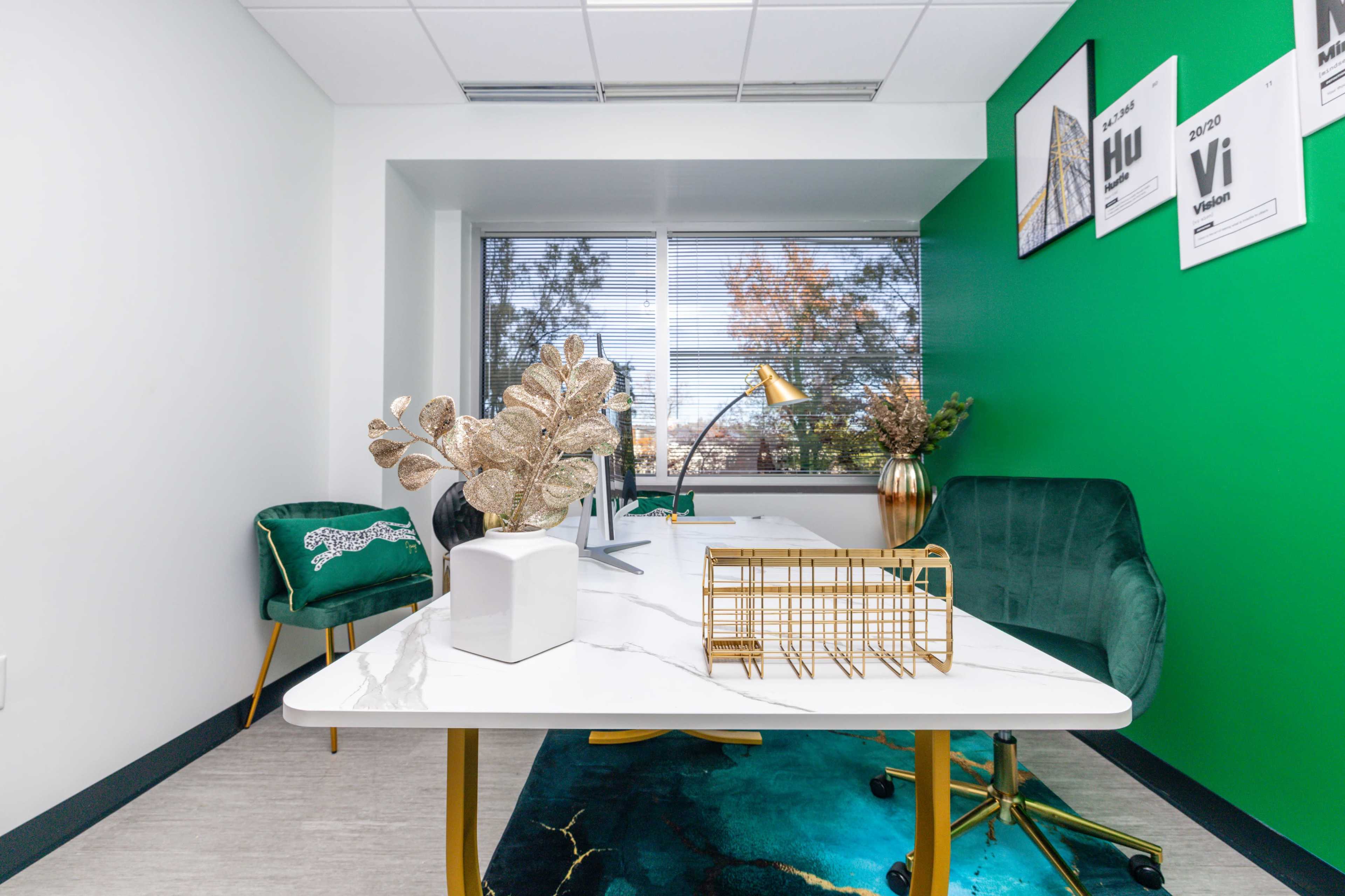 A modern office space with a green accent wall, a white desk, a gold desk lamp, and decorative plants near a large window.