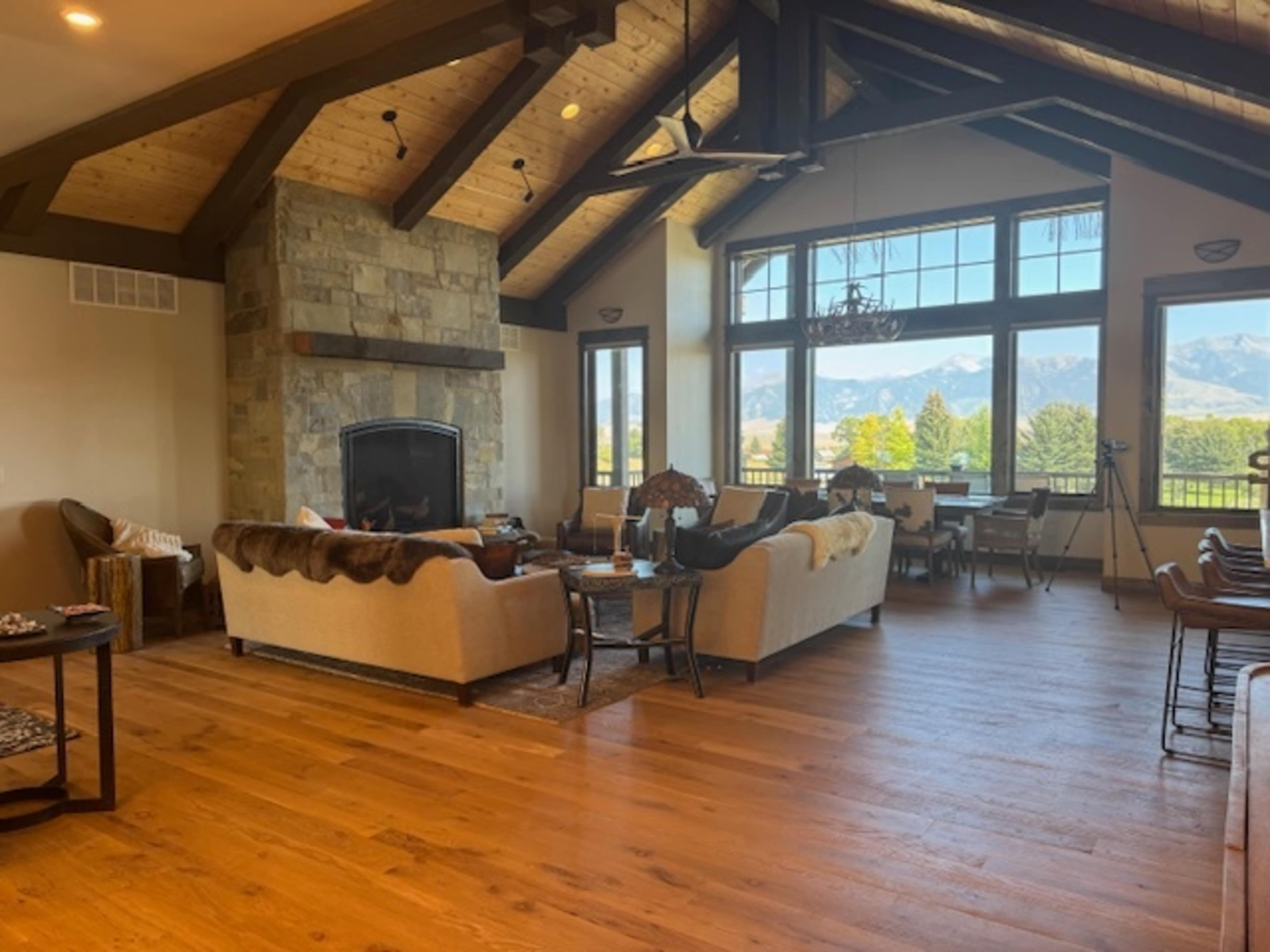 The image shows a spacious living room with a large stone fireplace, wooden beams, plush sofas, and large windows overlooking a mountainous landscape.