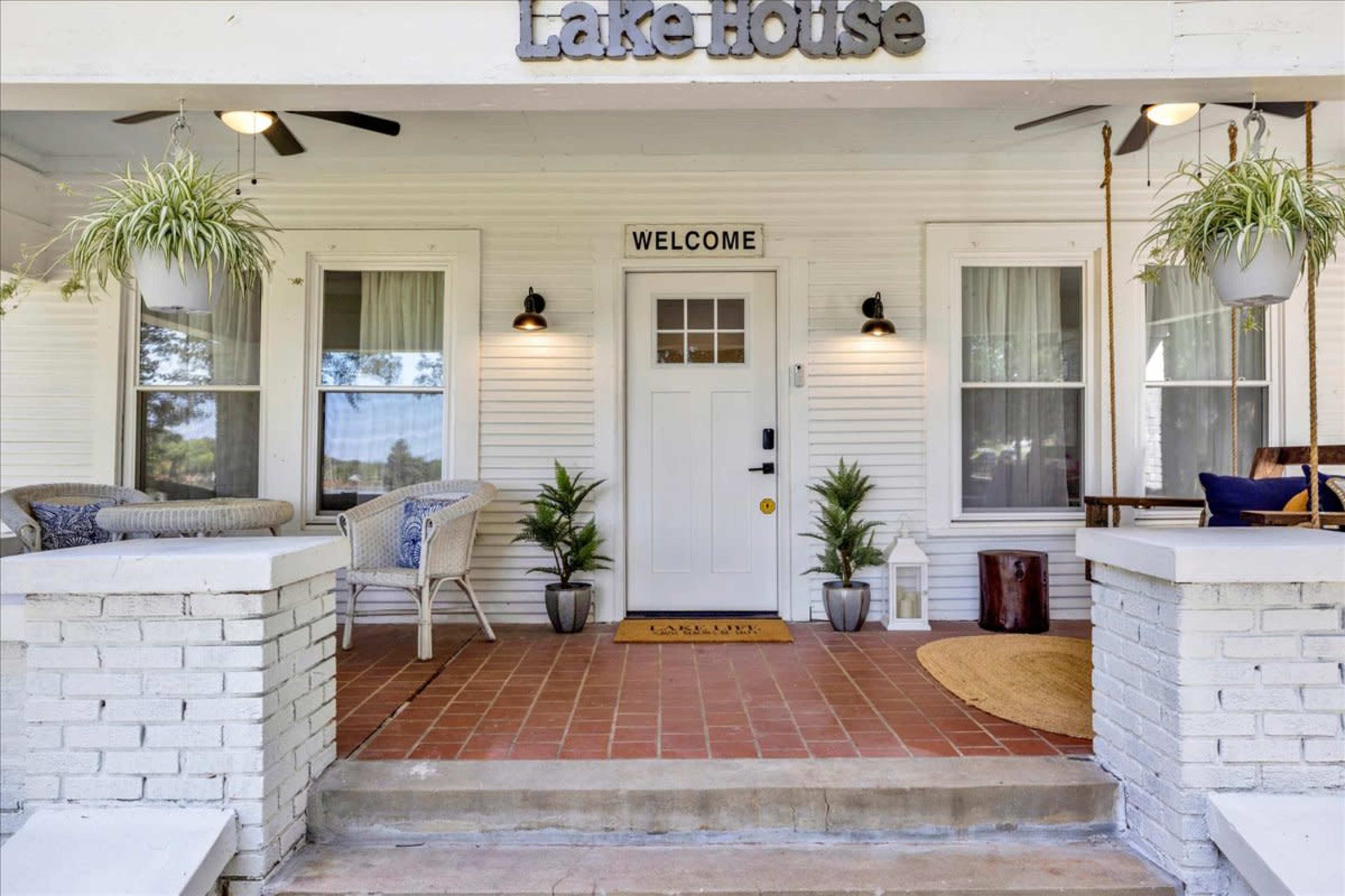 The image shows the front porch of a house, featuring a welcome mat, potted plants, and seating areas.