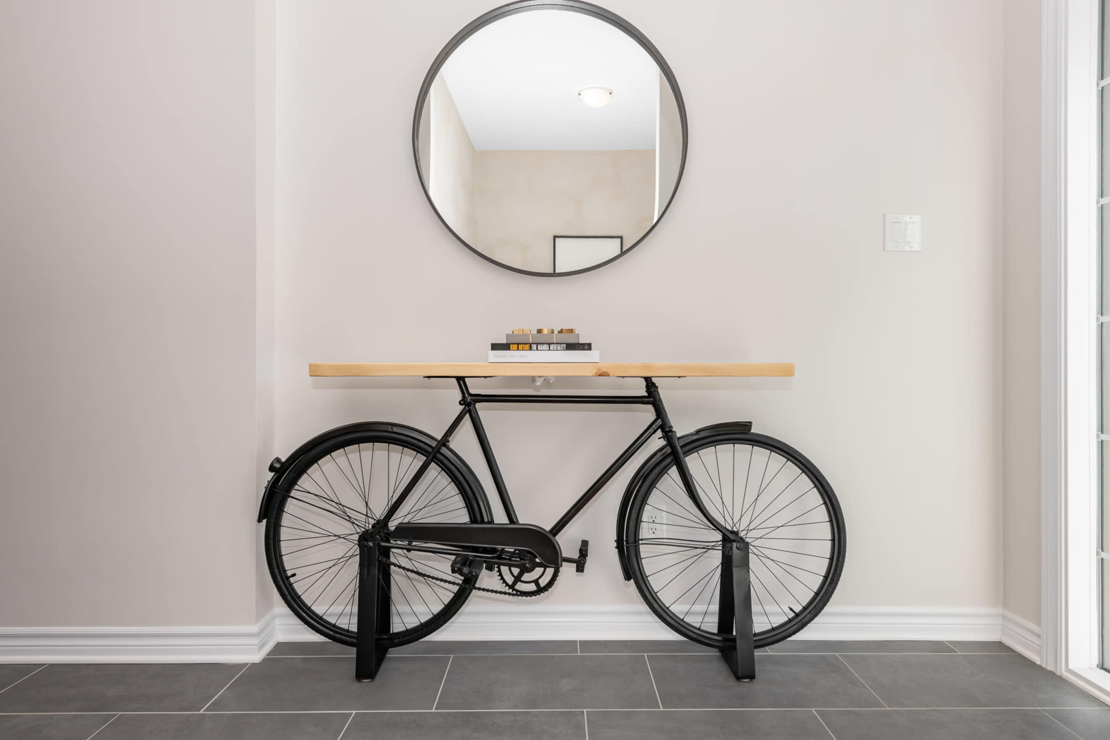 A black bicycle is positioned horizontally beneath a wooden tabletop, which is supported by a circular mirror on the wall above it.