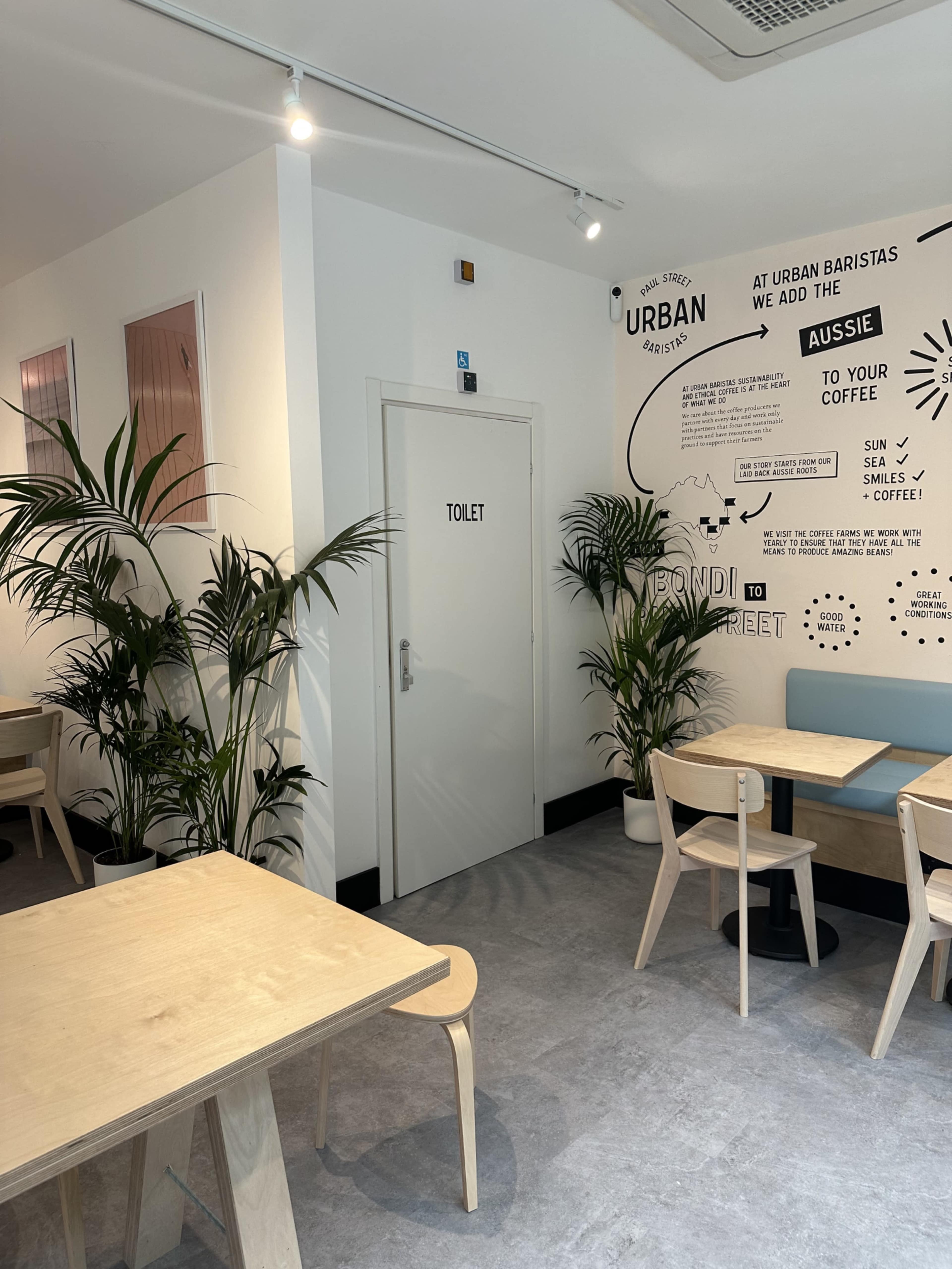 The image shows a modern café interior with light wood tables, green plants, and a door labeled "TOILET" on the wall, accompanied by decorative wall graphics.