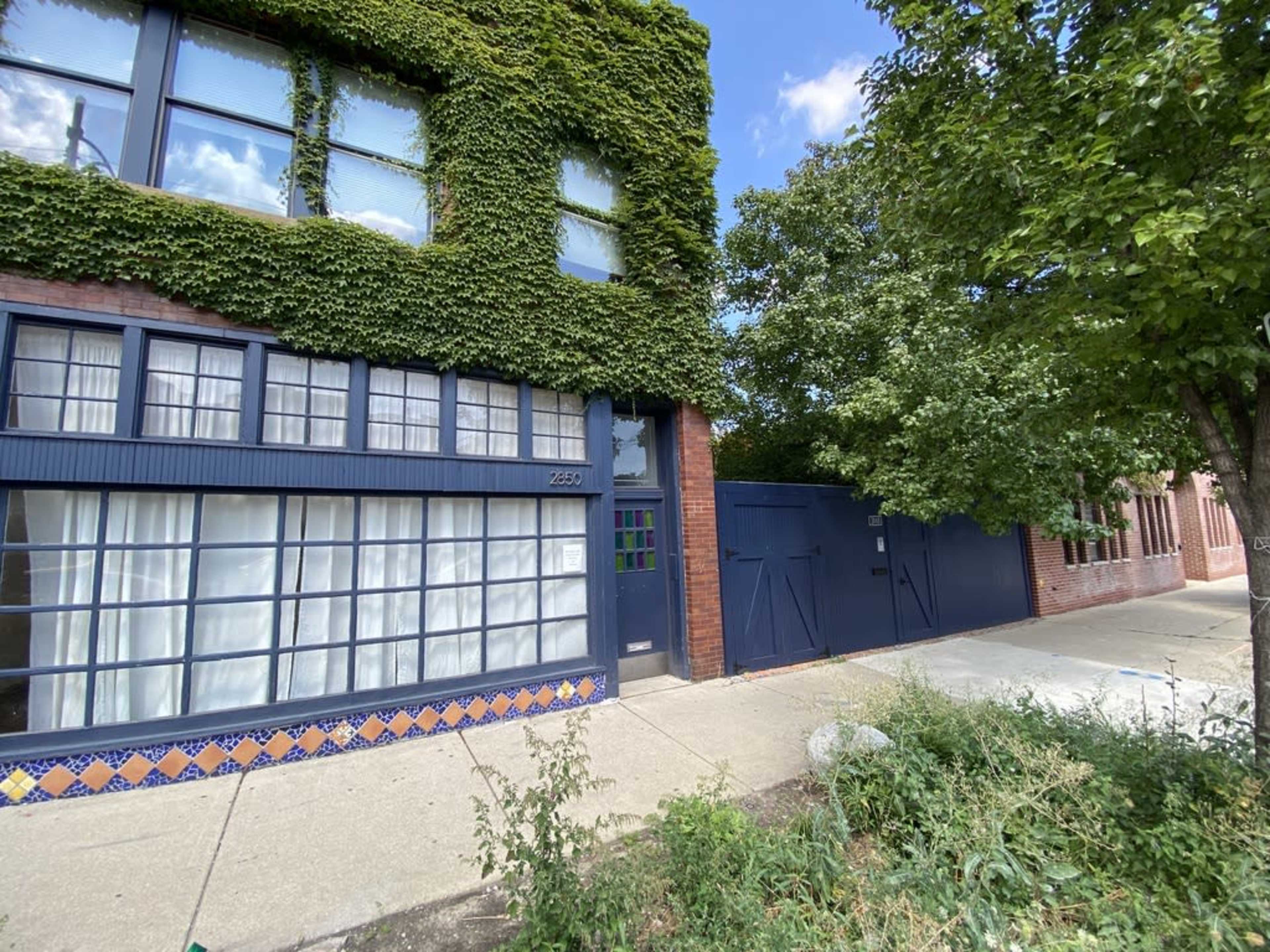 A building with an ivy-covered exterior features large windows and a blue door, set beside a paved walkway and greenery.