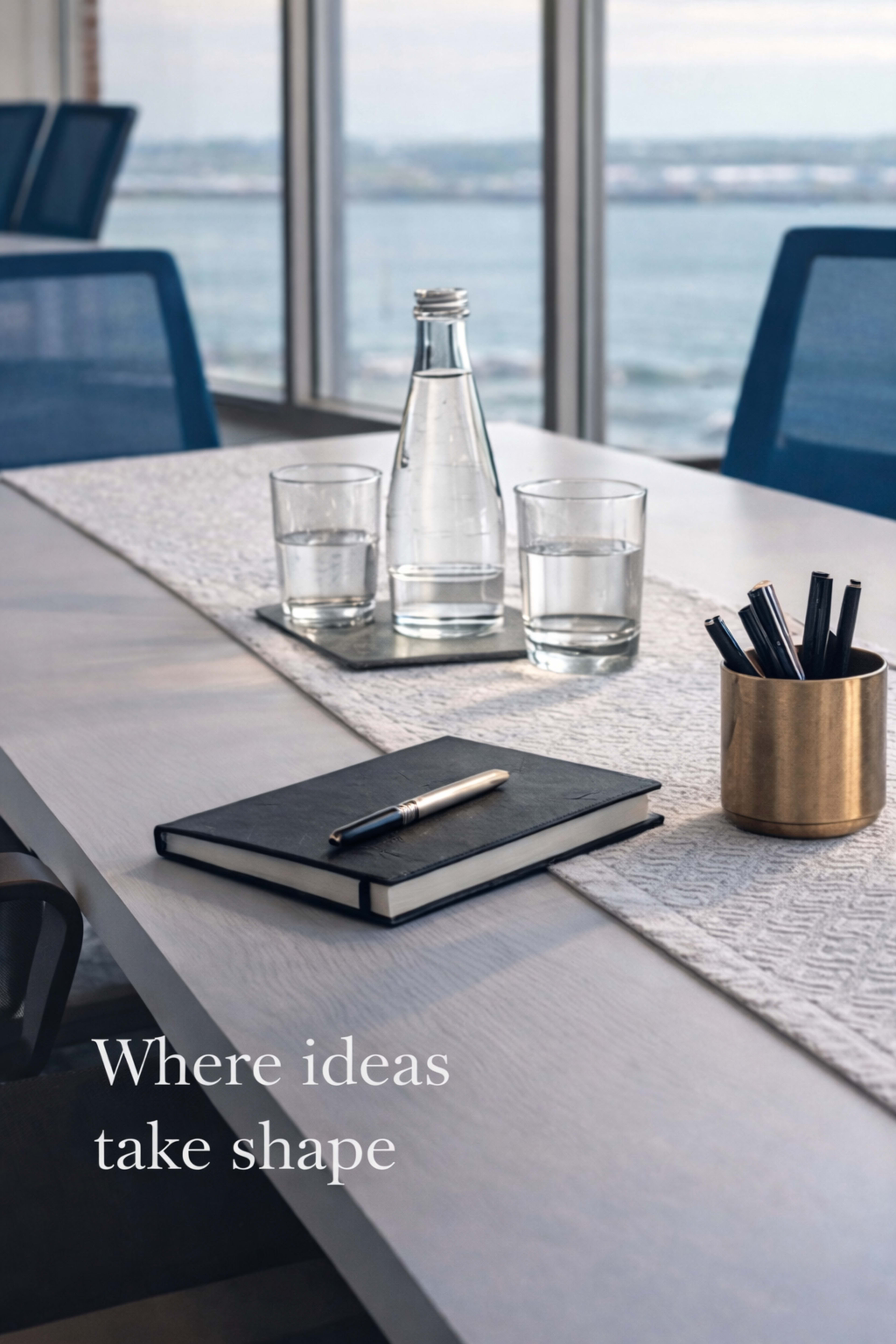 A conference table is set with a notebook, a pen, a glass water bottle, and two glasses of water, overlooking a scenic view of water and sky.